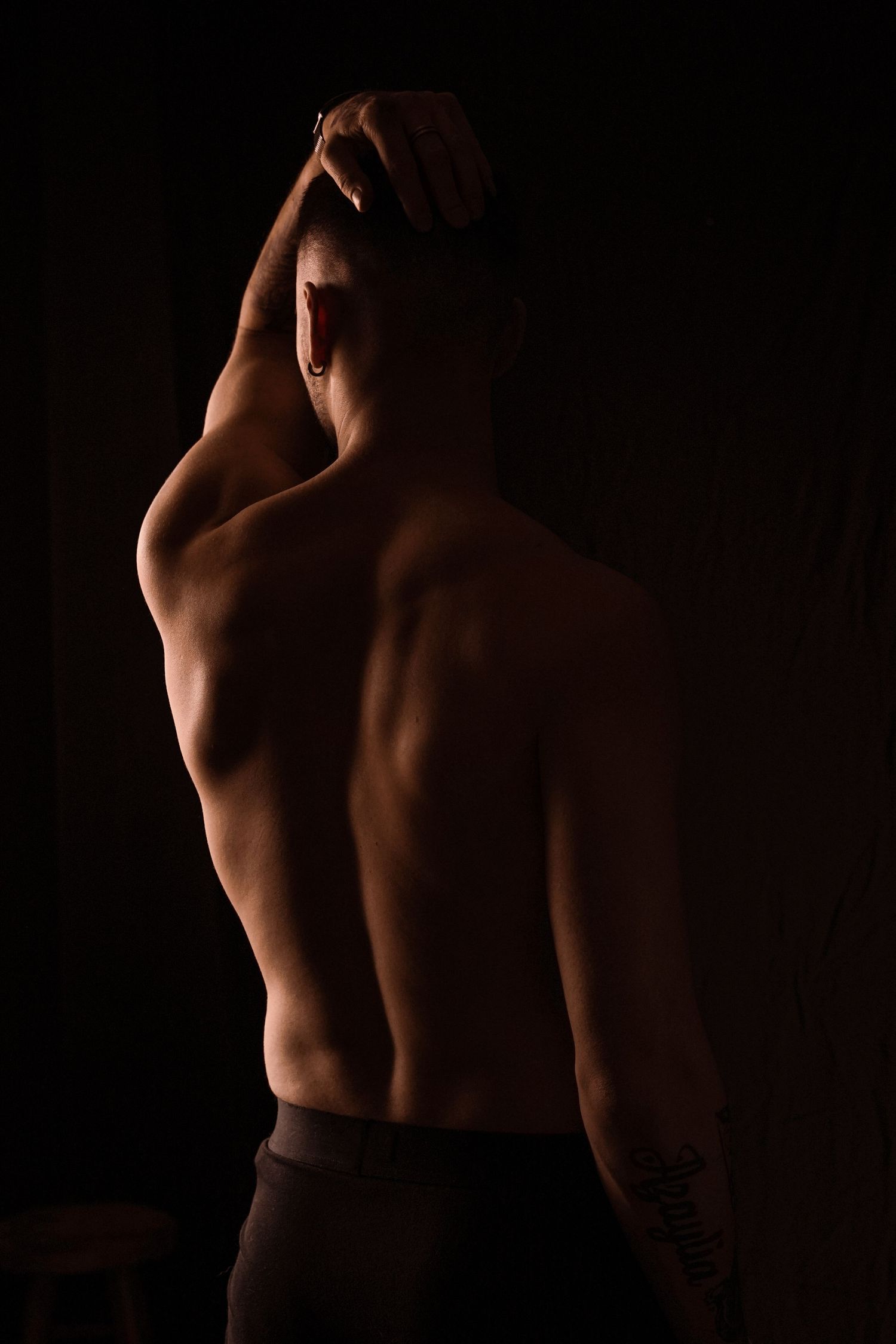 Shirtless muscular man viewed from behind with hand raised to head in dark dramatic lighting.