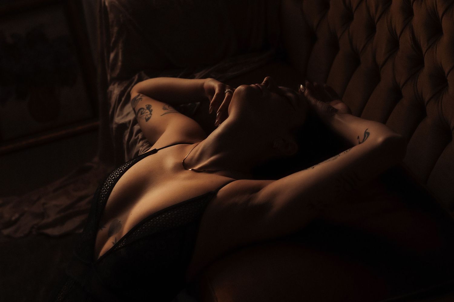 A woman in dark lingerie reclines on a tufted sofa in atmospheric moody boudoir photography.