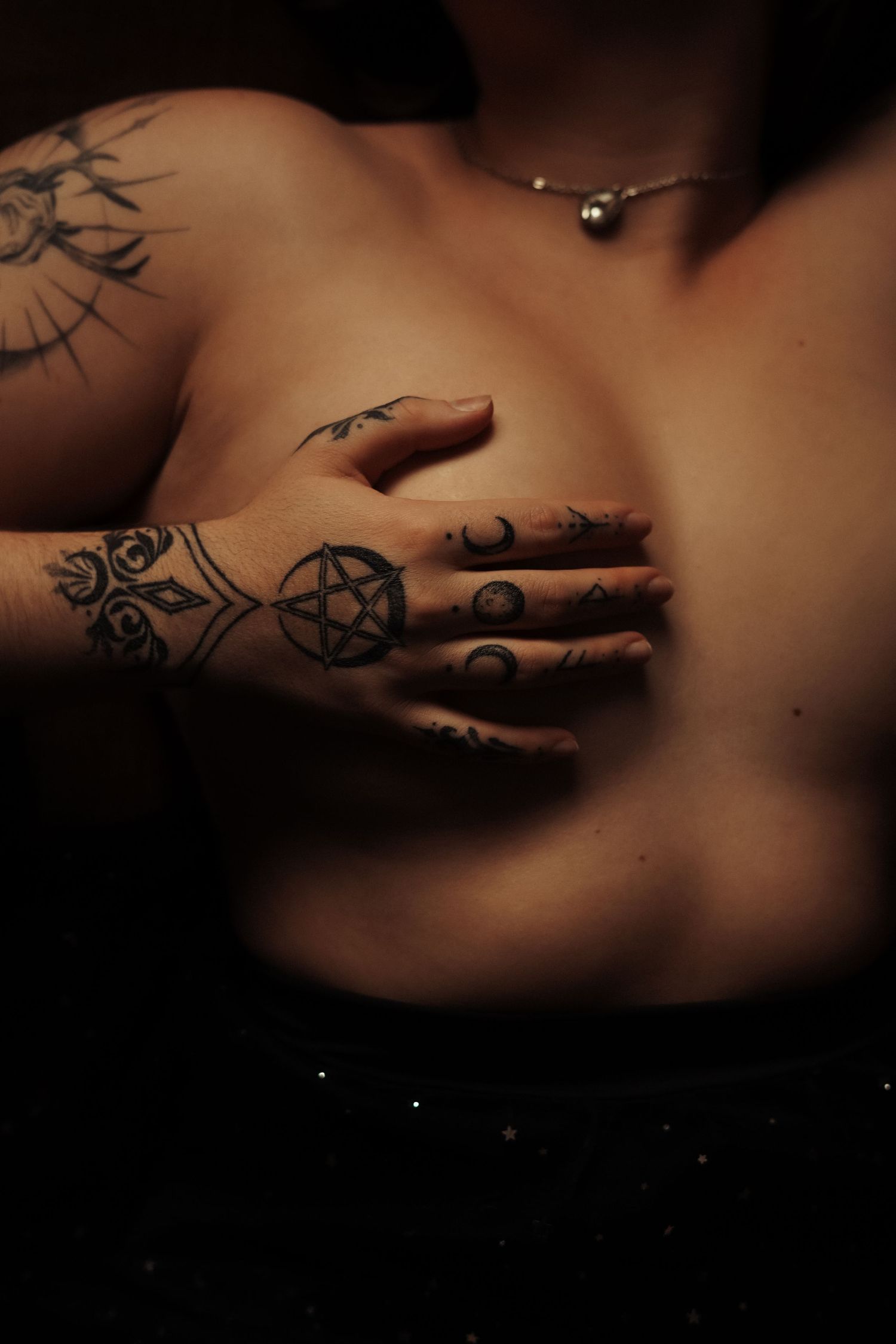 Close-up of tattooed hand with pentagram design covering chest in dark mystical boudoir photography.