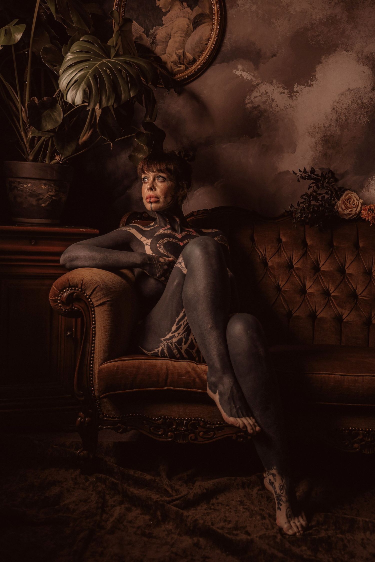 Tattooed woman in black bodysuit posing on velvet chair in dark moody studio.