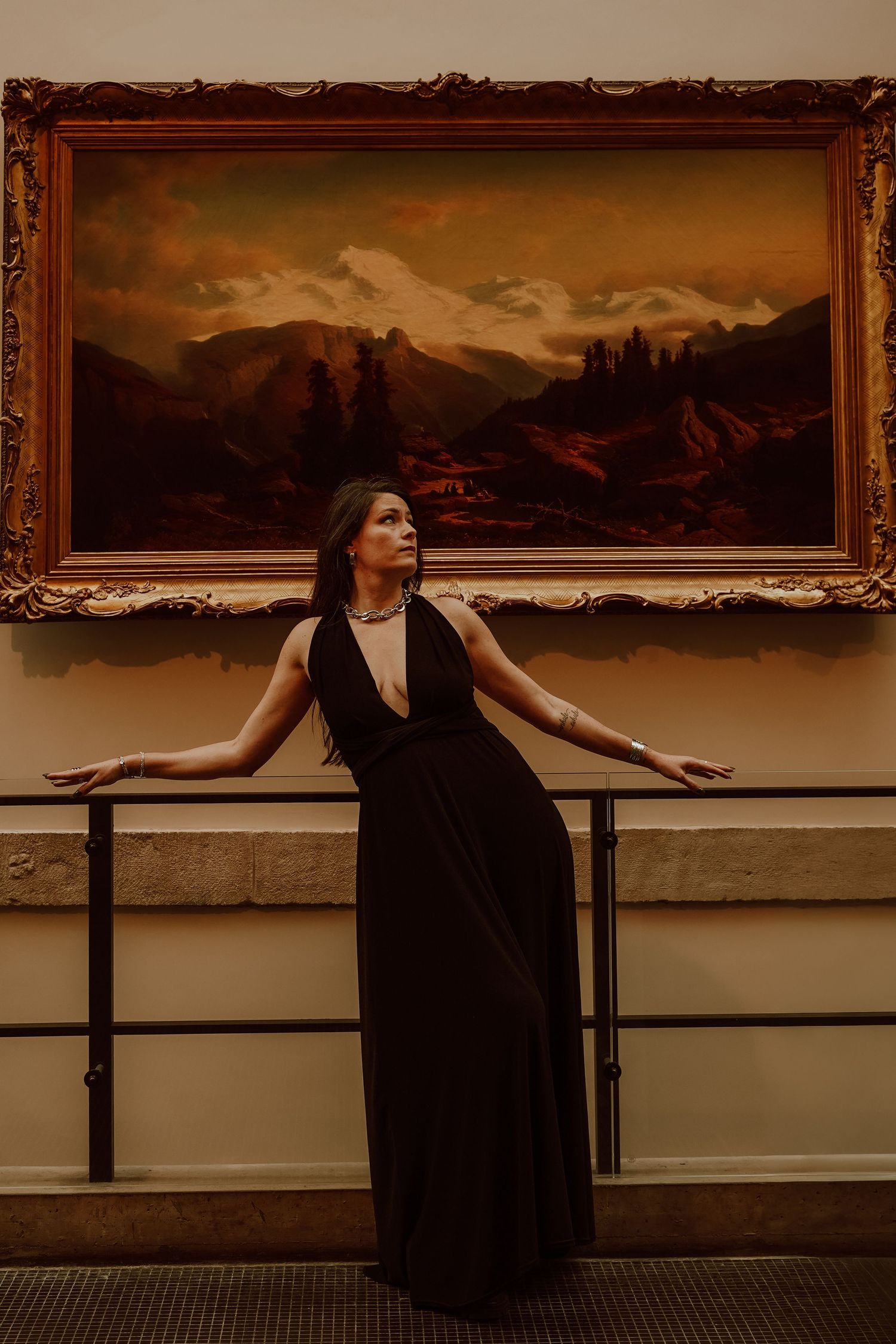 Woman in black gown poses elegantly before a framed mountain landscape painting in a gallery.