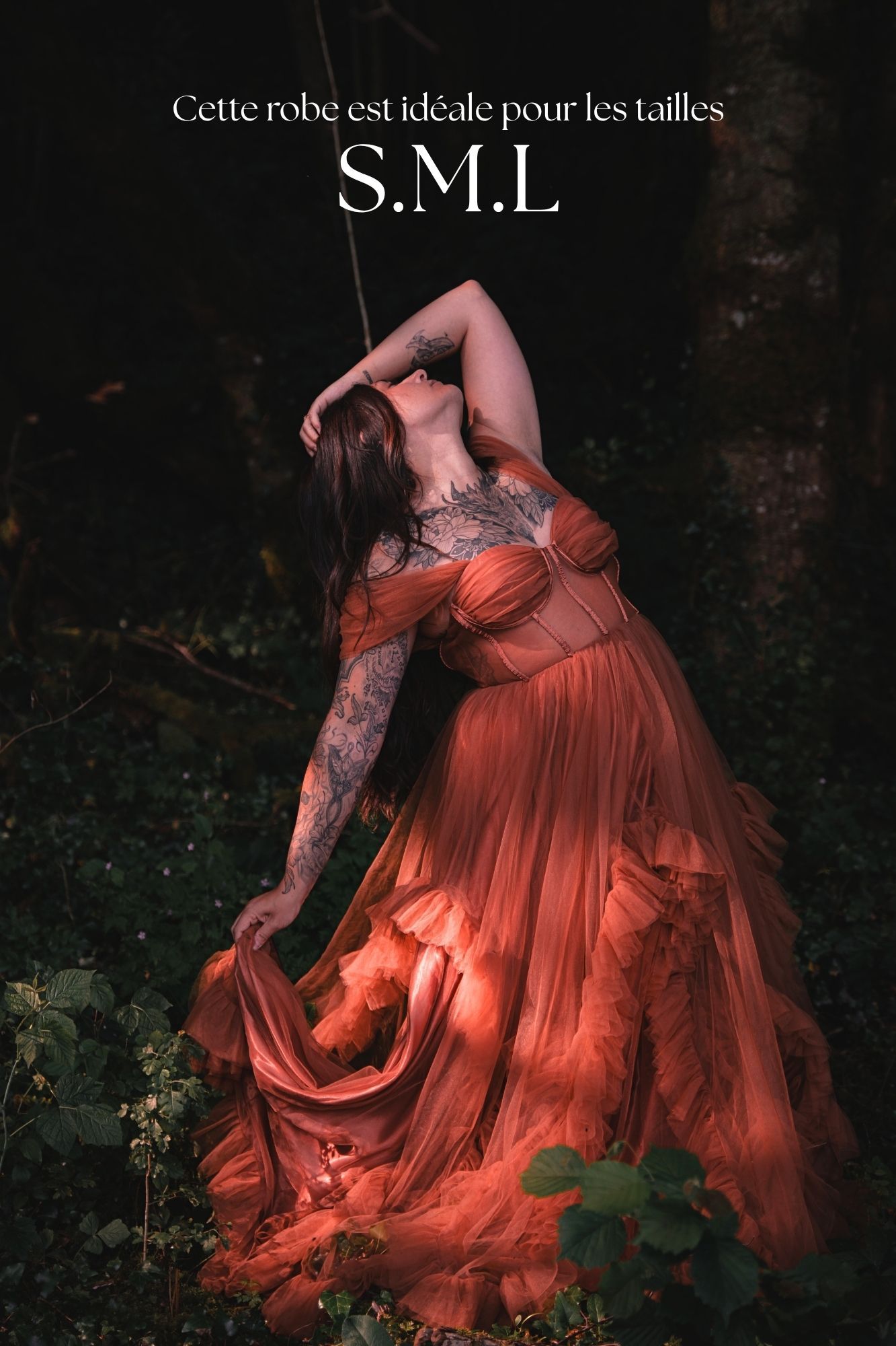 A woman in a flowing rust-orange tulle gown poses dramatically in a dark forest, ideal for sizes S, M, and L.