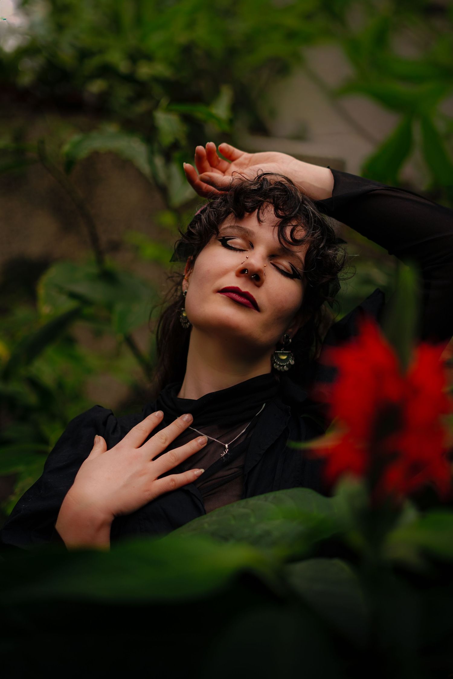 Woman in black outfit with eyes closed posing among red flowers in green garden.