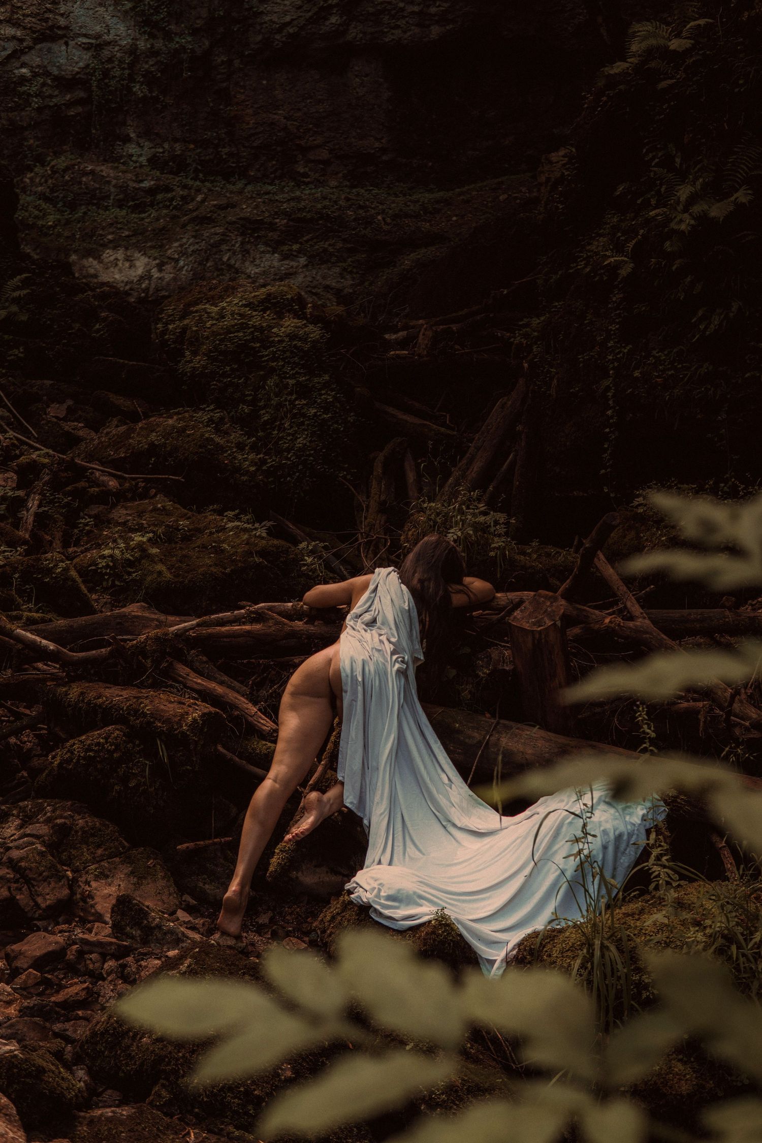 Woman in flowing white sheer fabric posed among mossy rocks and roots in a dark enchanted forest.
