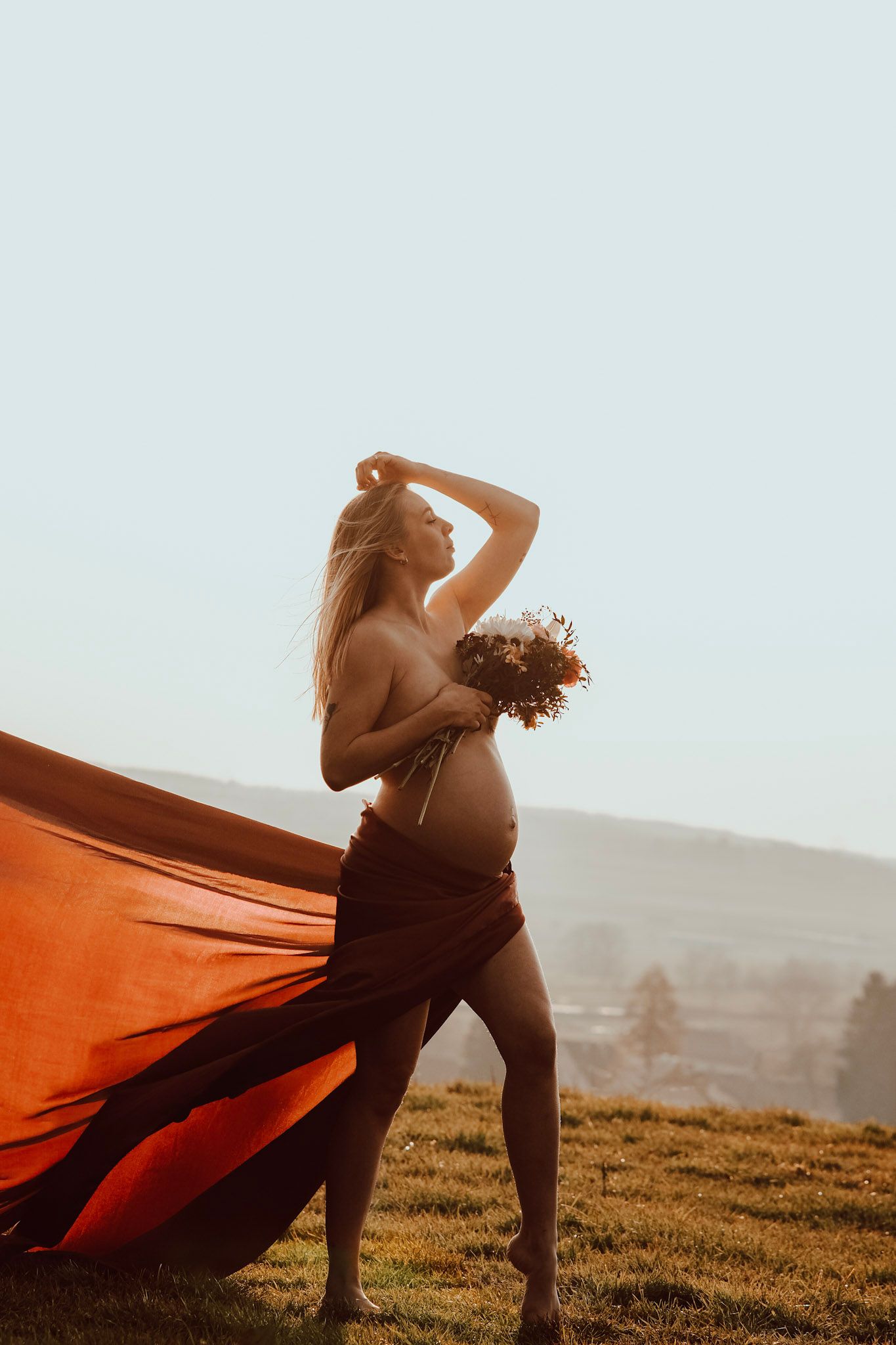 Pregnant woman in flowing orange fabric holding wildflowers outdoors at golden hour.