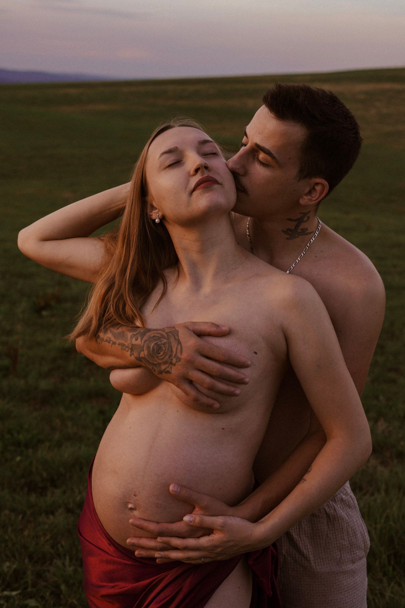 Couple sharing intimate moment outdoors at dusk, pregnant tattooed woman in red fabric.