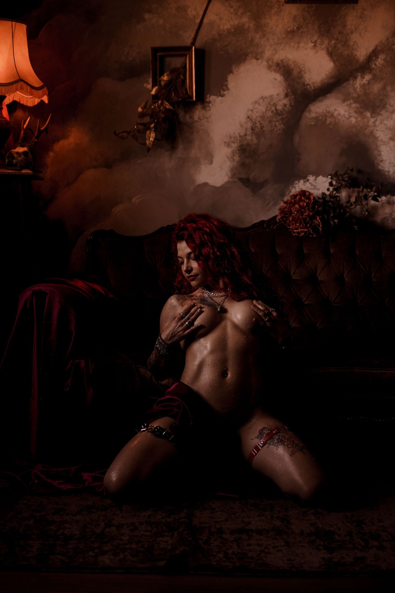 Red-haired tattooed woman posing topless in dramatic moody dark room setting.