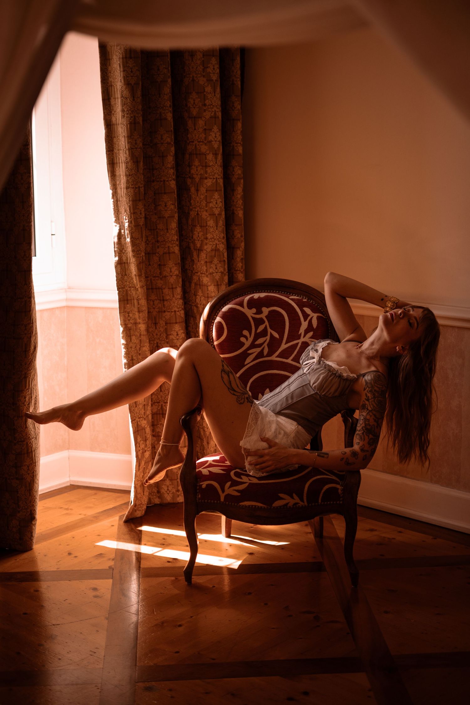 Woman in lingerie posing dramatically in antique chair by sunlit window on hardwood floor.