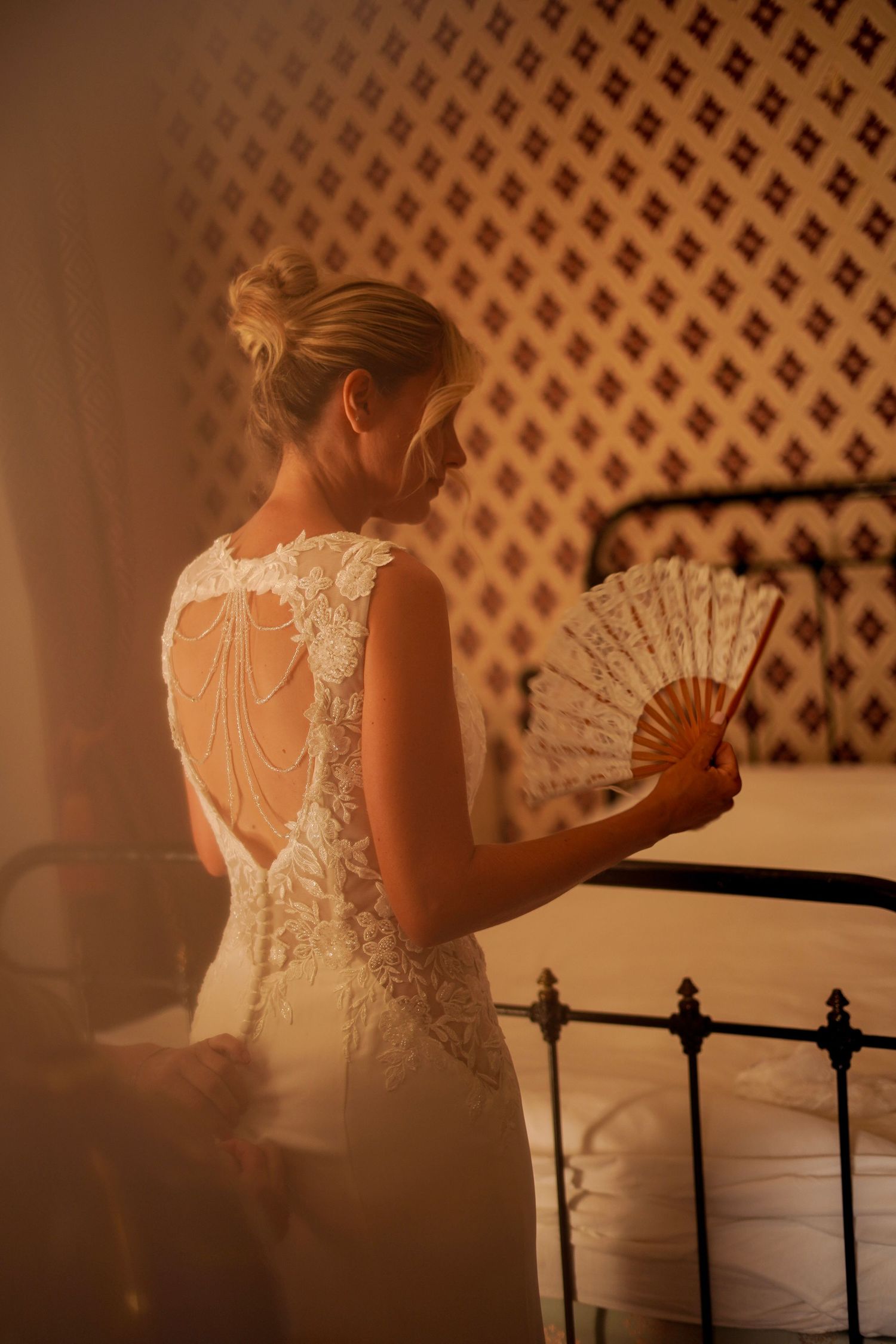 Bride in lace wedding dress holding decorative fan, showcasing elegant pearl back detail in warm golden light.