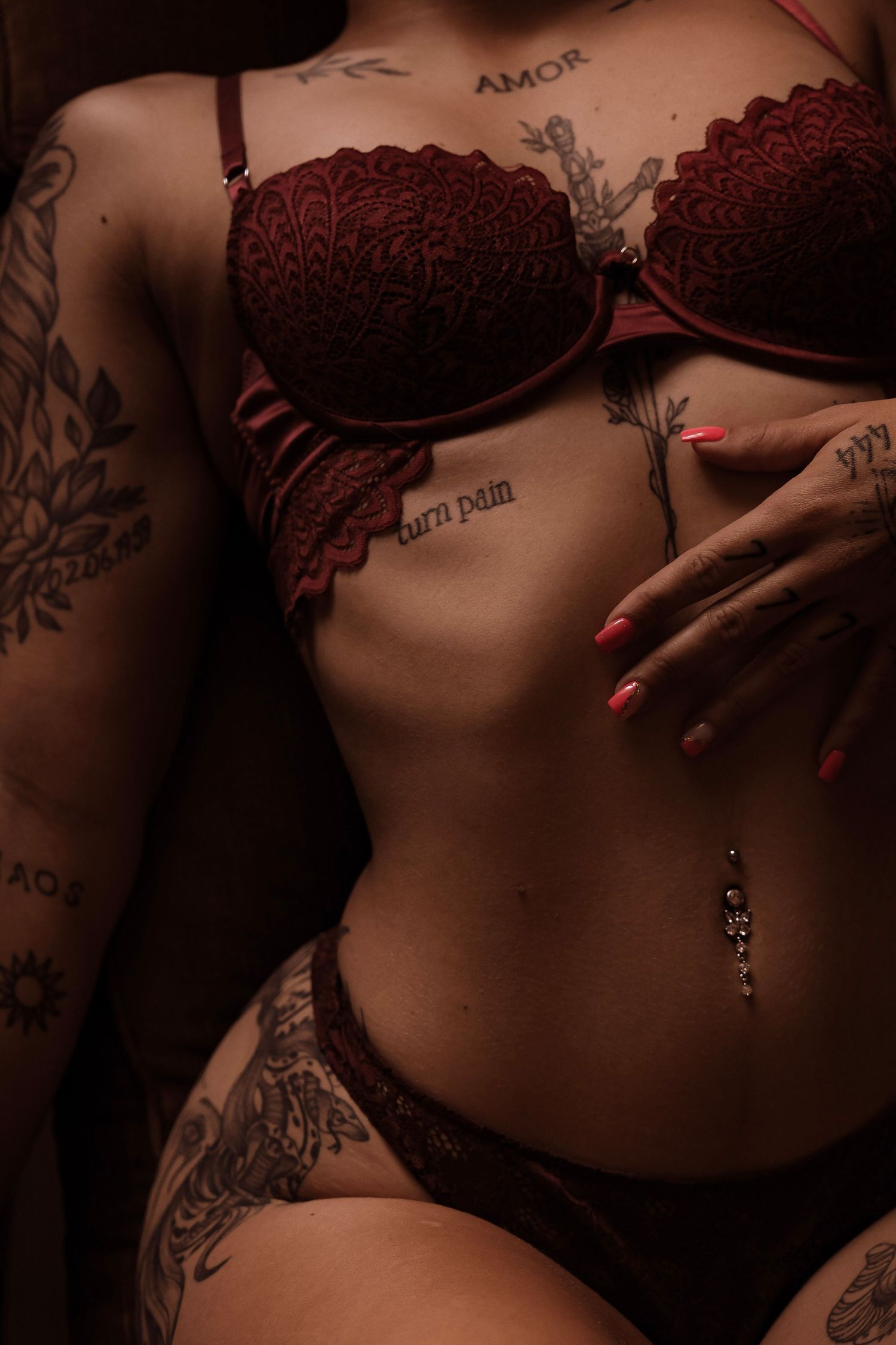 Tattooed woman in red lace lingerie set with belly piercing and red nails.