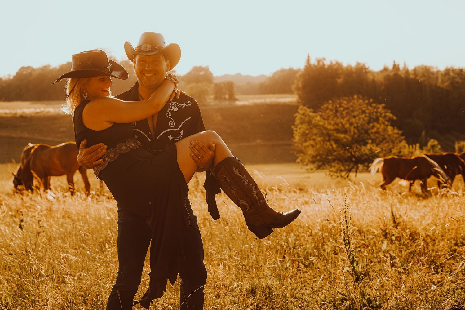 A couple in cowboy hats shares a romantic sunset moment in a golden field with horses grazing behind.