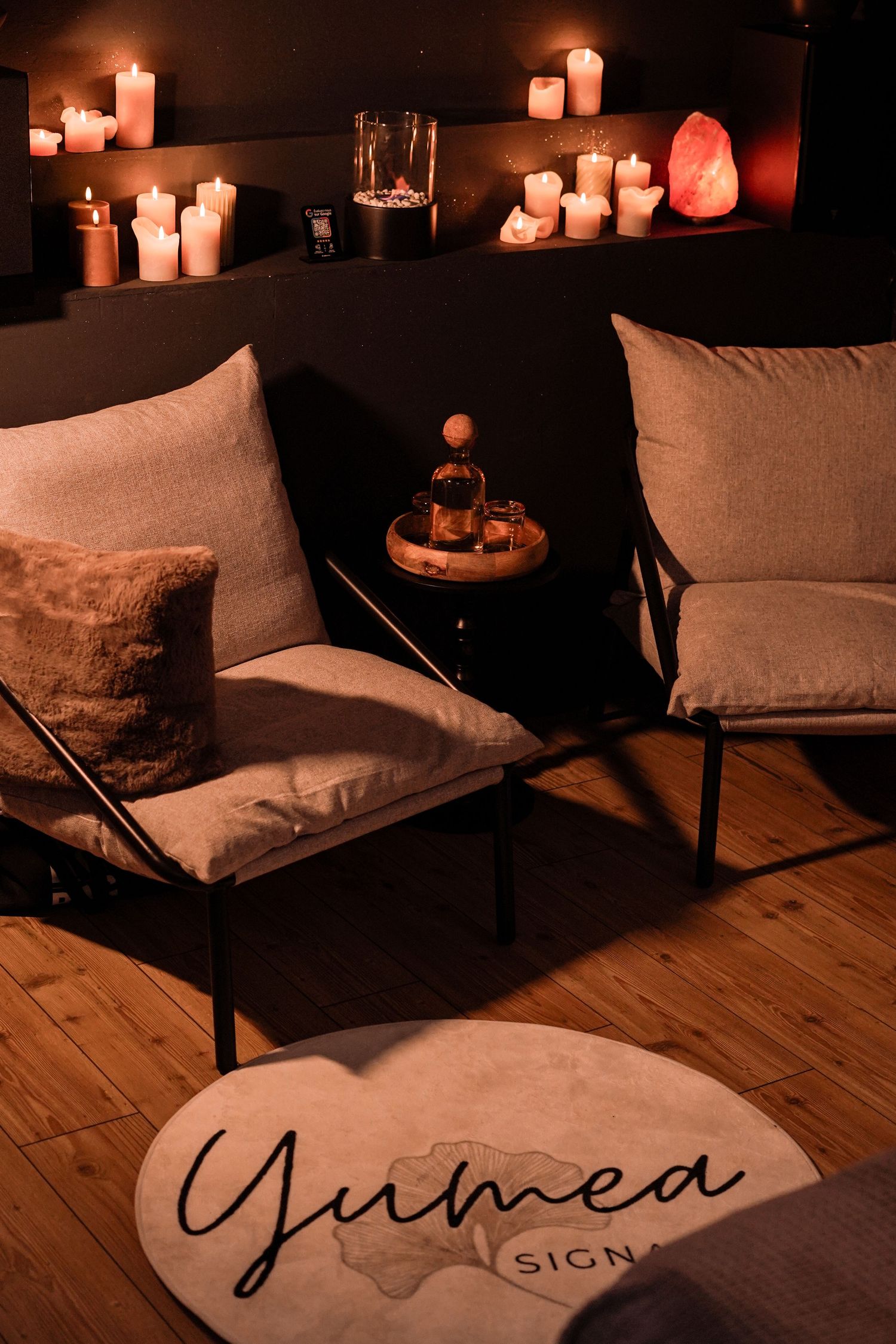 Cozy candlelit spa lounge with gray chairs, pillows, and Yumea Signature branded round rug.