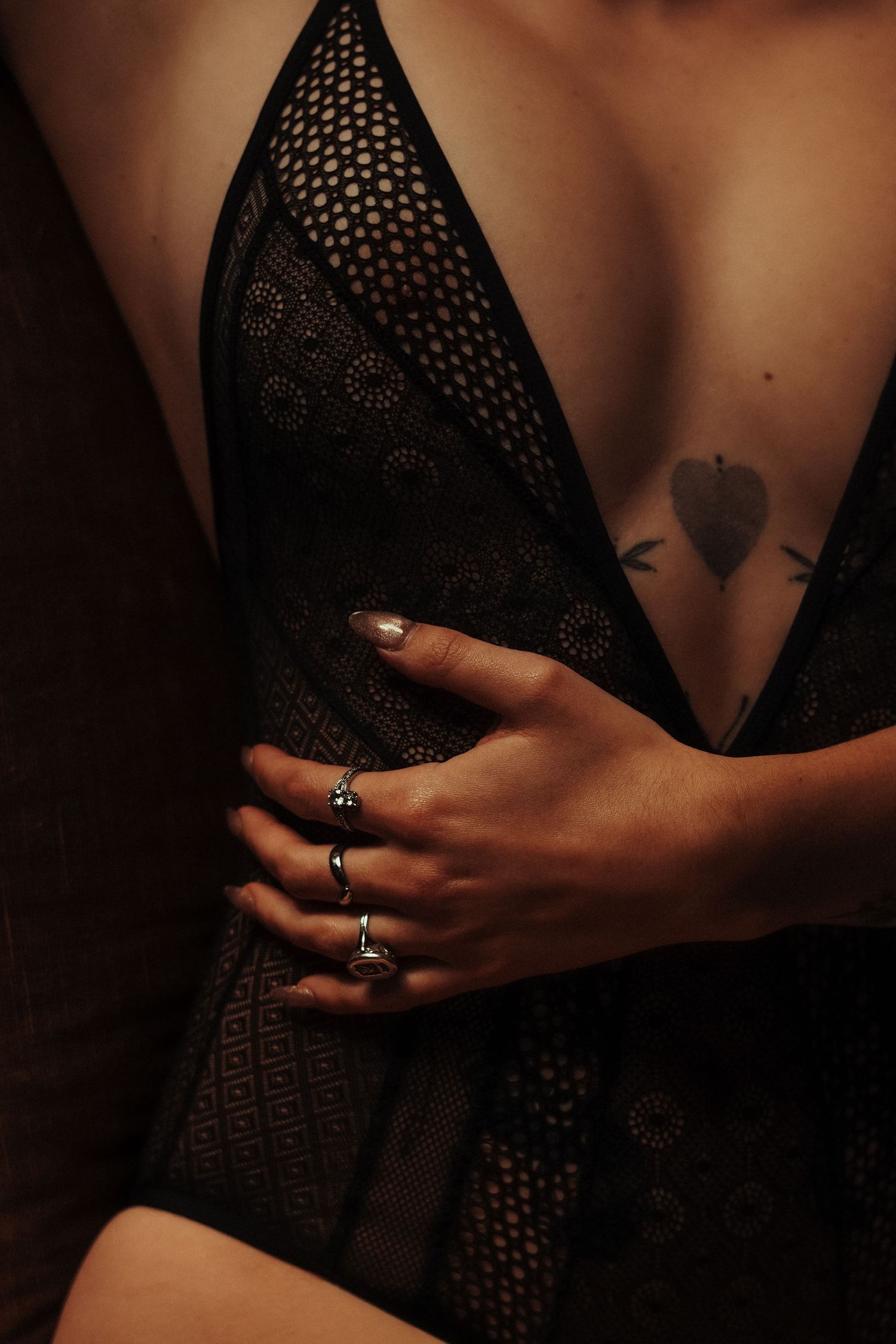 Woman in black lace bodysuit with heart tattoo and rings posing in moody low-light boudoir photography.