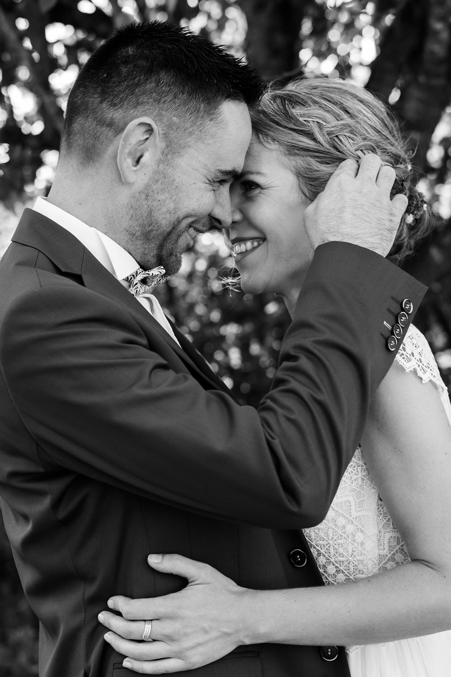 Black and white photo of smiling bride and groom embracing forehead to forehead outdoors.