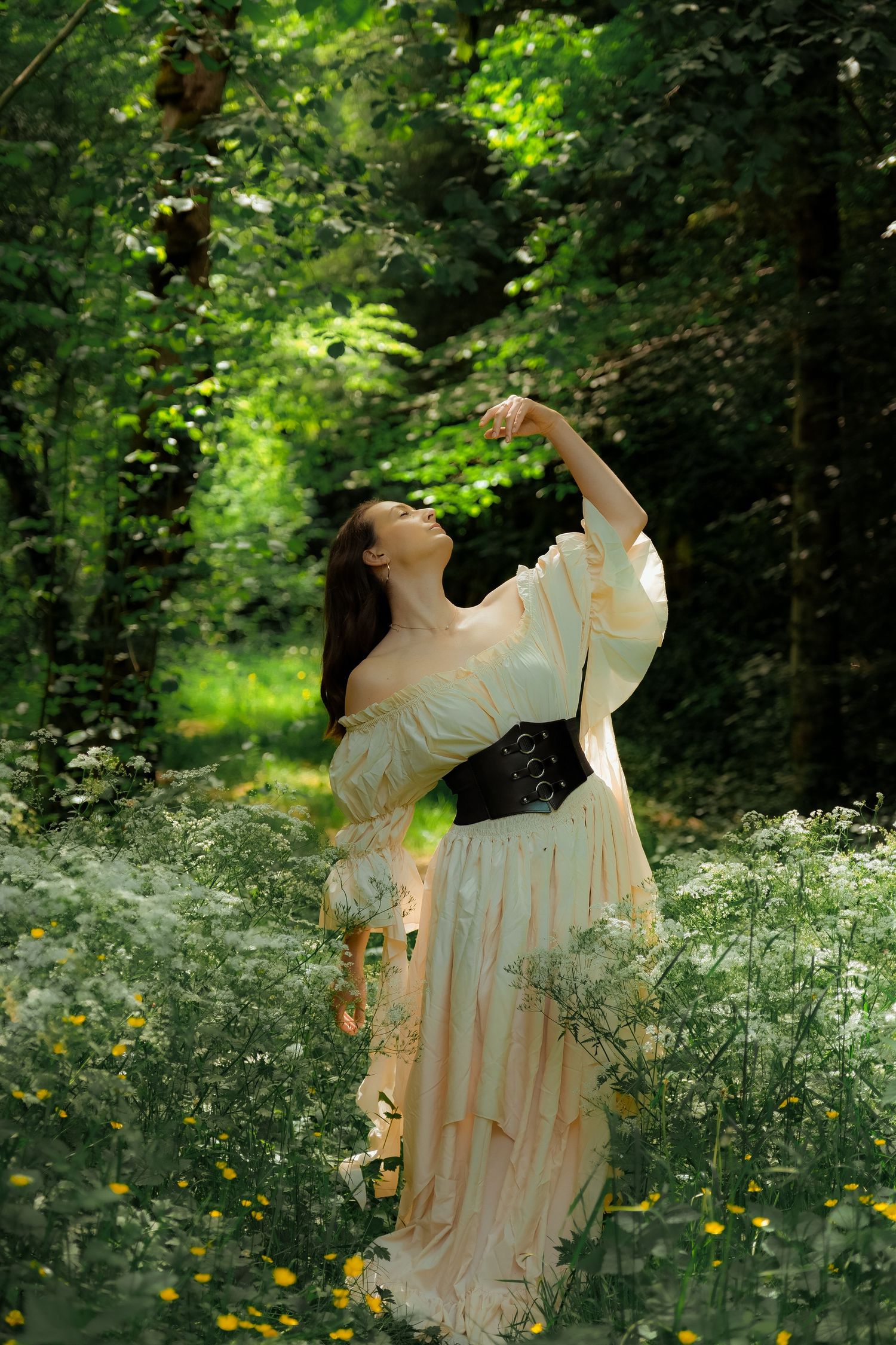 Woman in cream off-shoulder gown posing gracefully among wildflowers in a lush green forest.