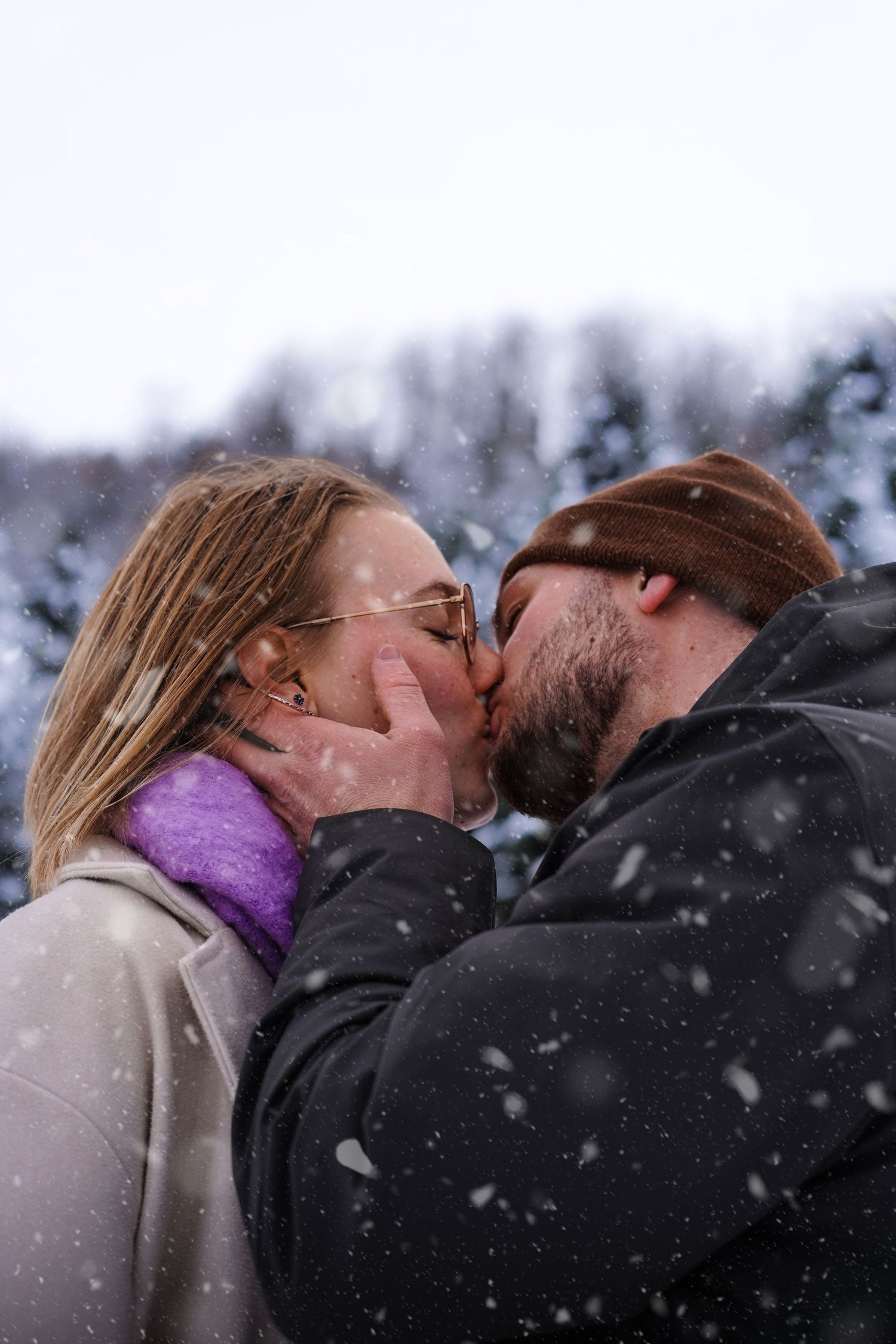 Couple sharing a kiss in falling snow wearing winter coats and accessories.