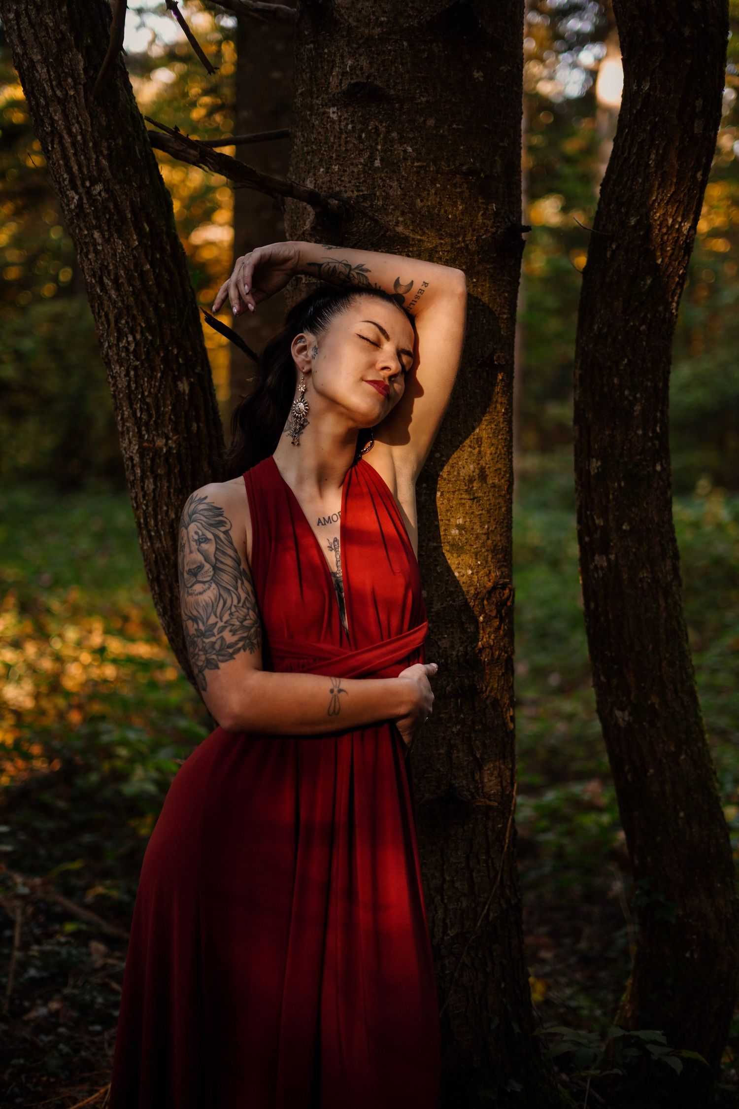 Tattooed woman in red dress leaning against trees in a sunlit autumn forest.