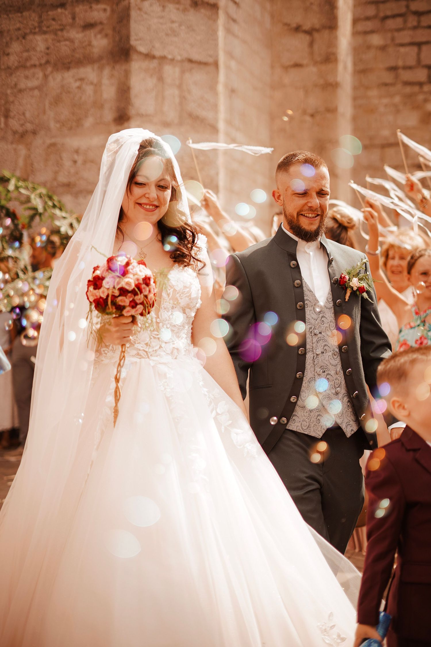 Happy bride and groom walking out of church surrounded by bubbles and cheering wedding guests.