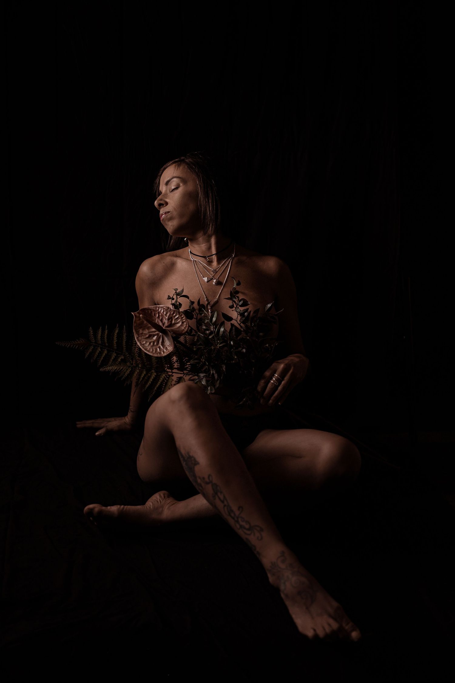 A woman poses gracefully with tropical flowers against a dark background in a fine art boudoir photo.
