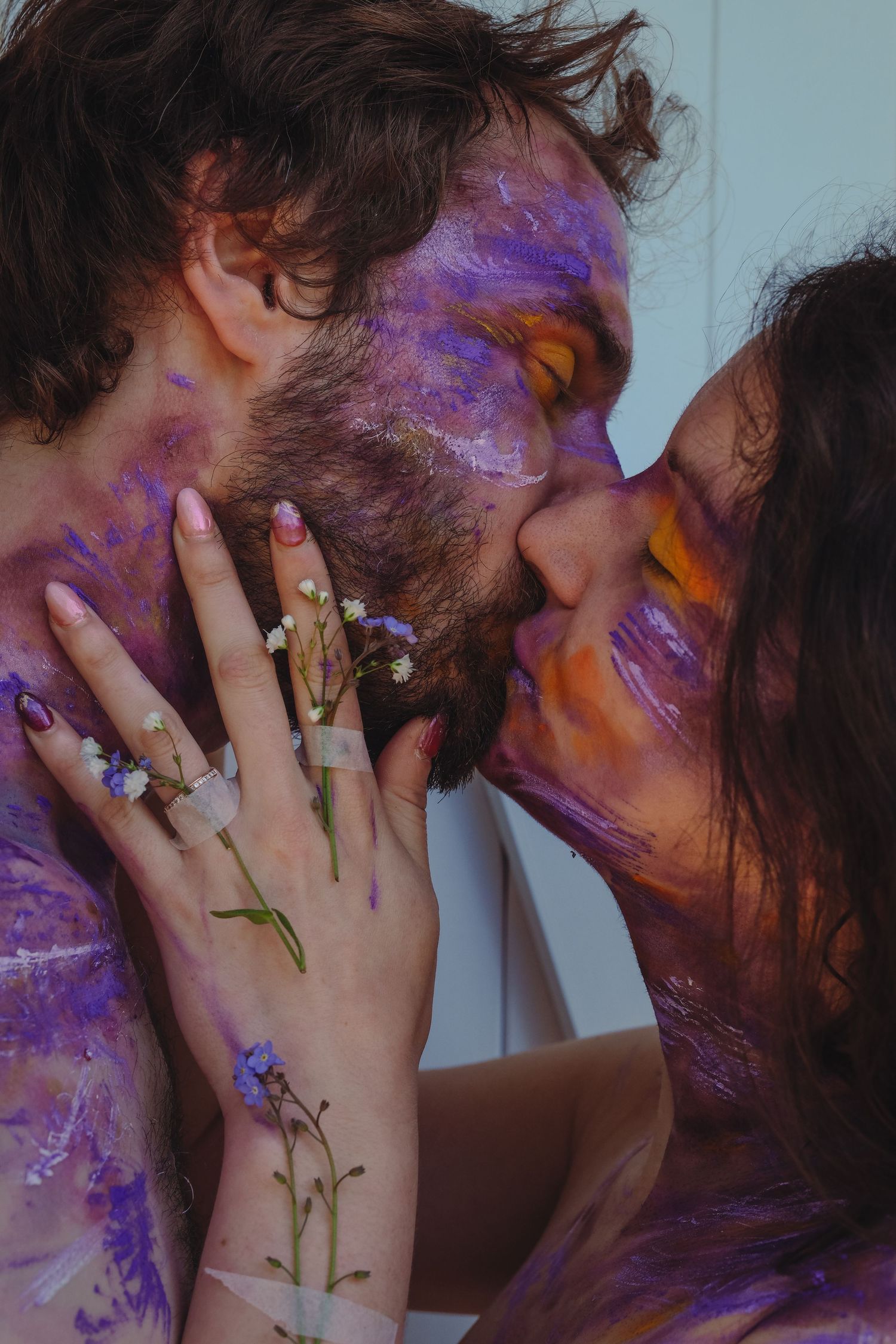 Couple with purple and yellow paint on faces sharing intimate kiss with floral accents.
