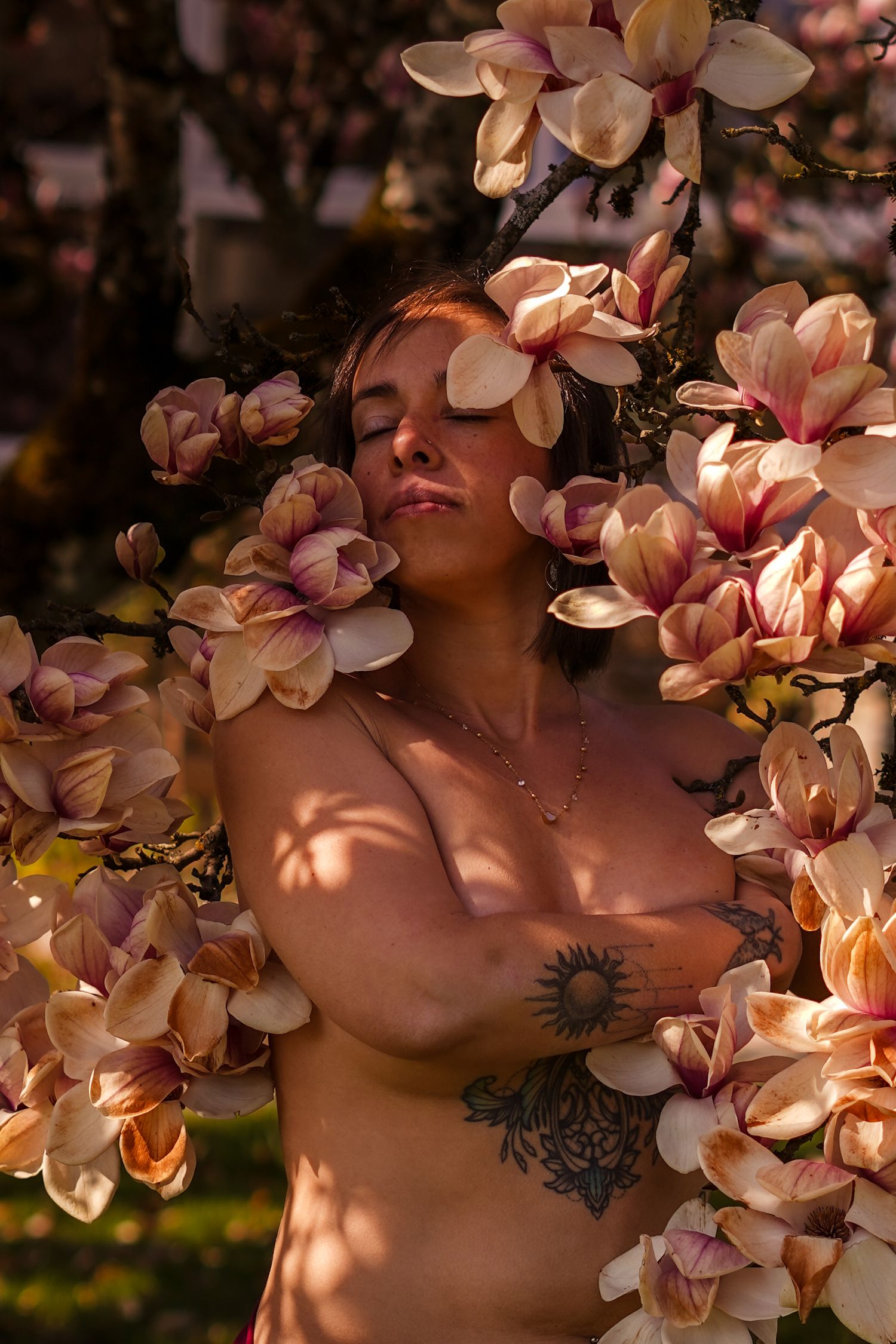 Woman with tattoos posing topless among blooming magnolia flowers in warm sunlight.