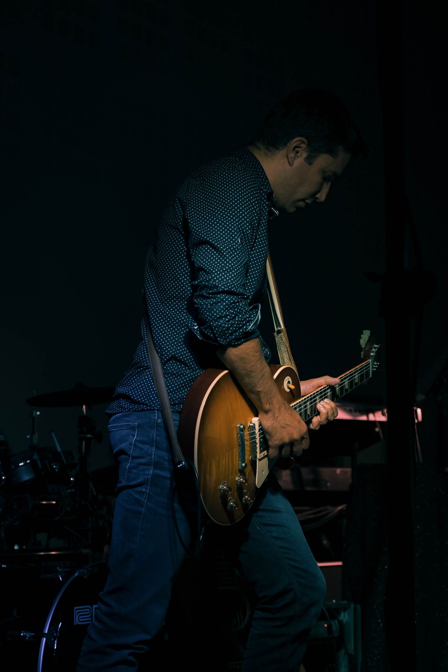 Male guitarist playing electric Les Paul guitar on dimly lit stage during live concert performance.