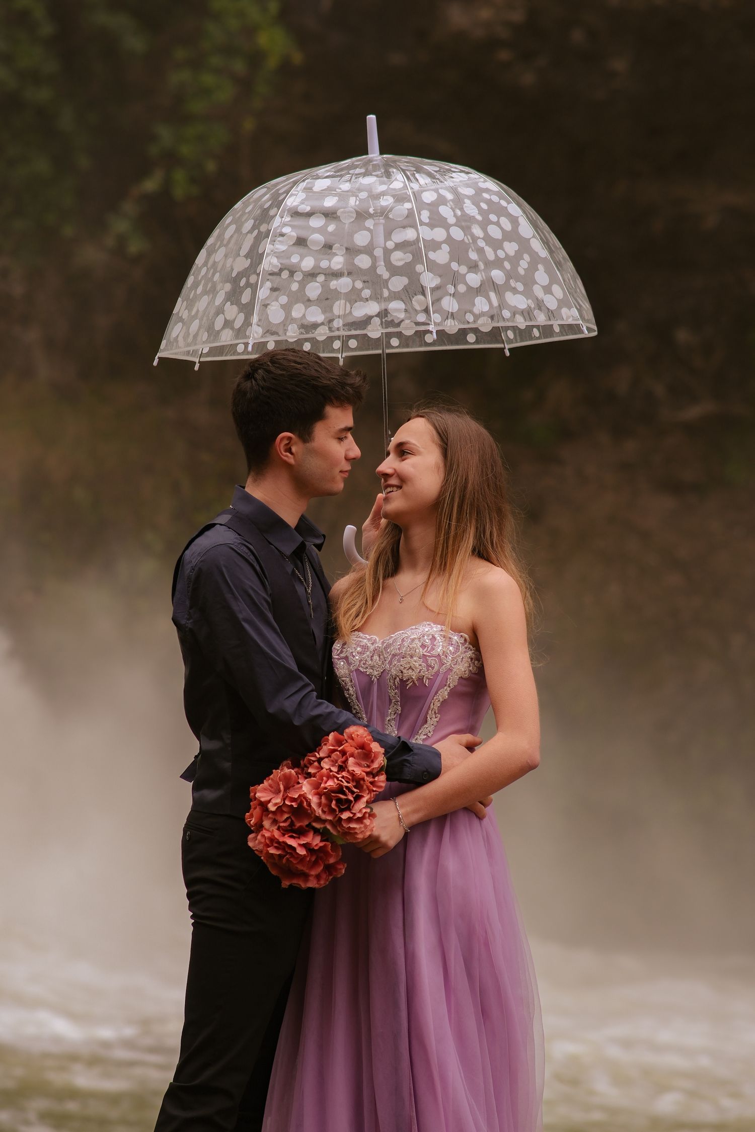 Elegant couple standing under a clear umbrella near a misty waterfall with red bouquet.