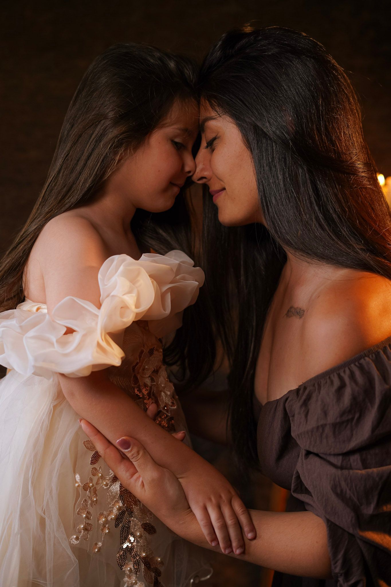 Mother and daughter touching foreheads tenderly in elegant dresses by candlelight.