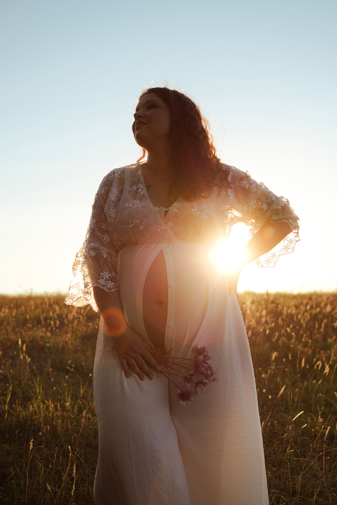 Pregnant woman in white lace dress standing in golden field with sun flare at sunset.
