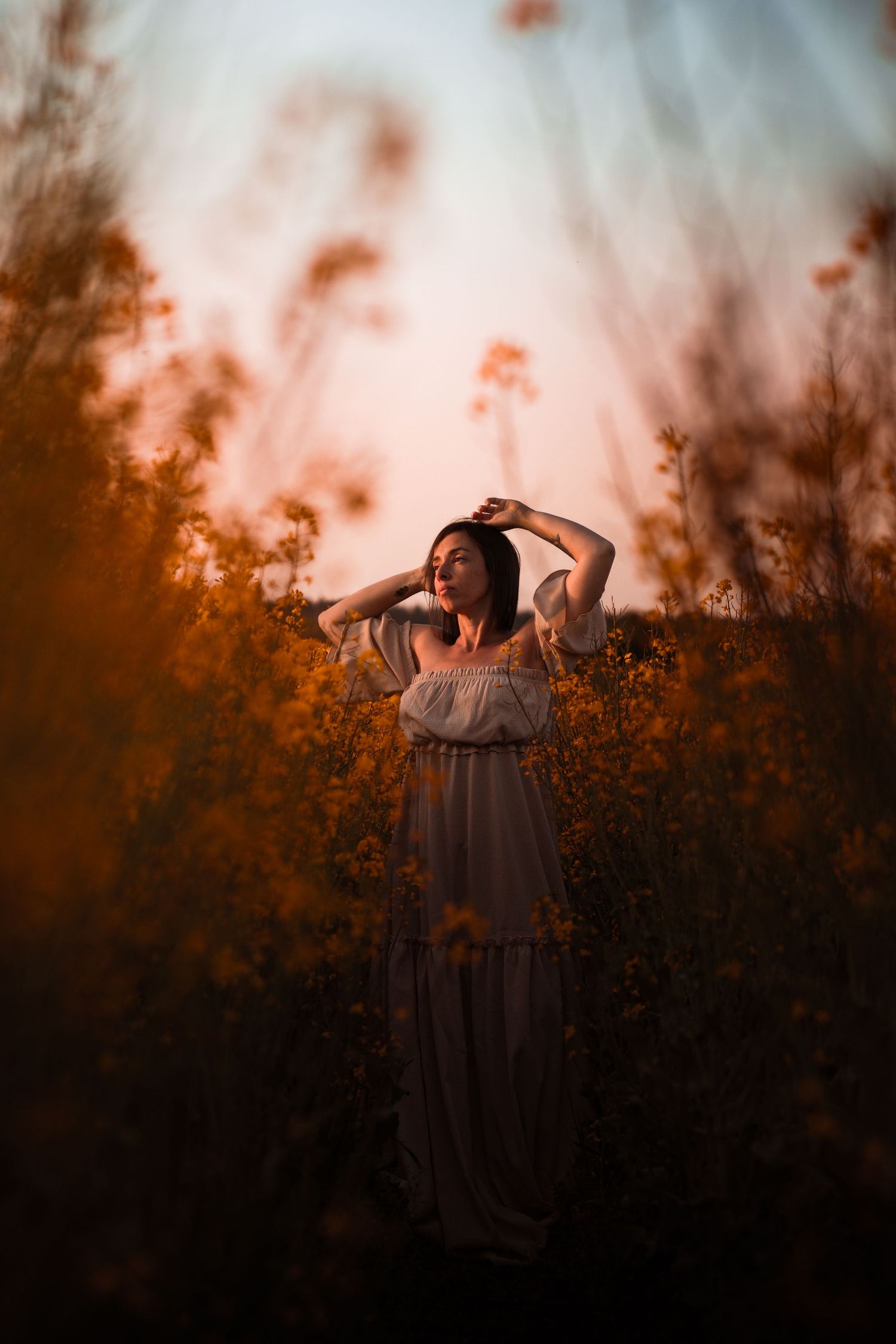 Woman in white off-shoulder dress stands among golden wildflowers in warm sunset light in open field.