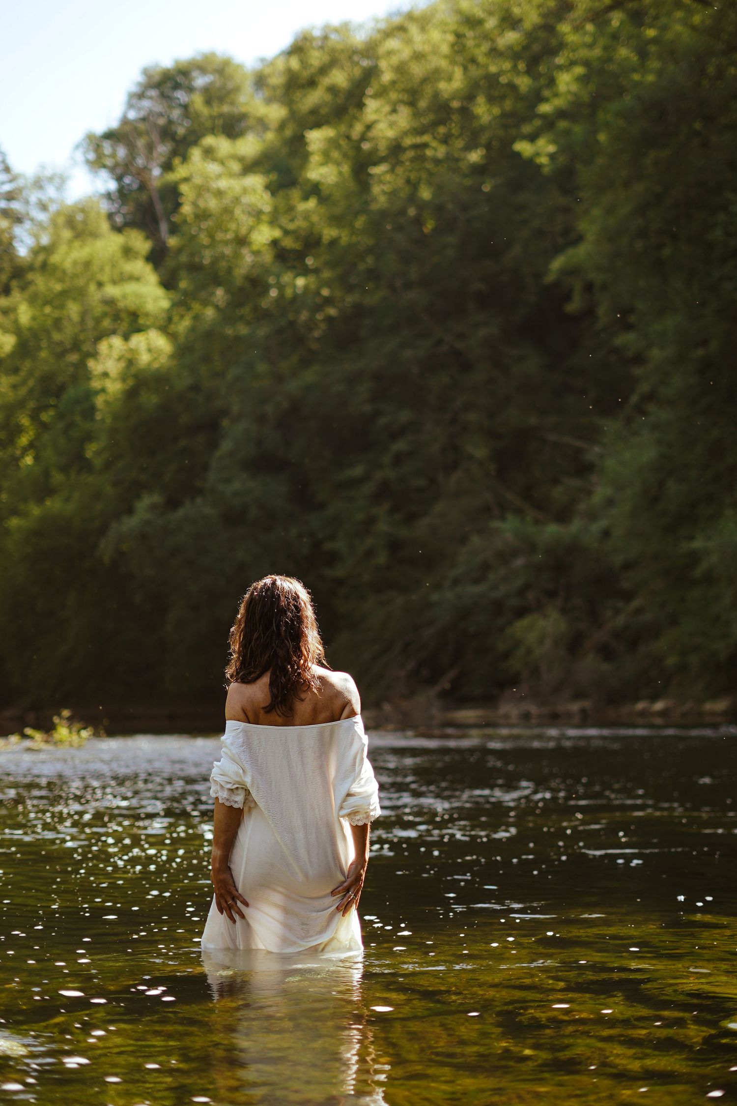 Woman in white dress wading in sunlit river with green forest backdrop.