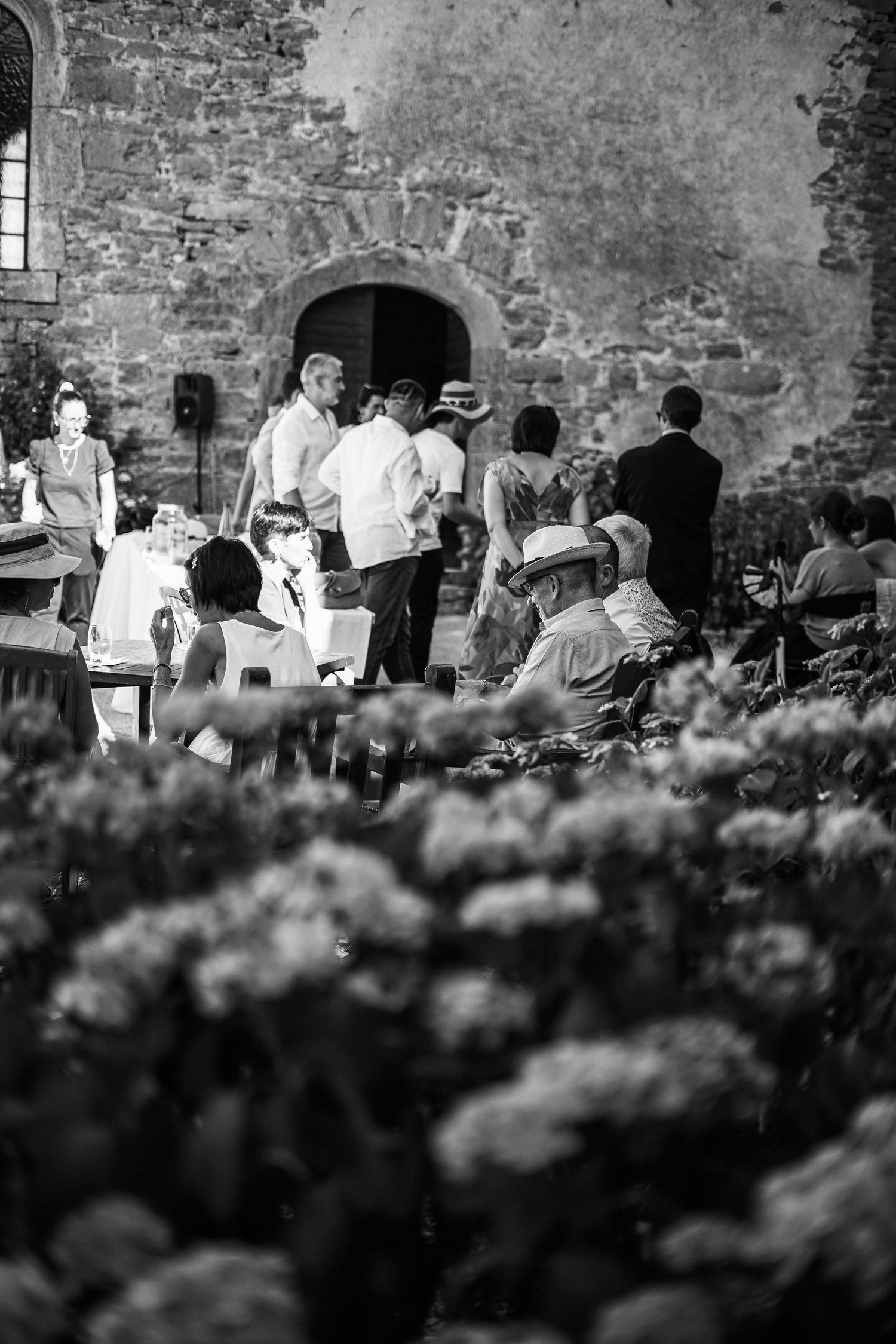 Black and white image of wedding guests gathered in stone courtyard surrounded by flowers.