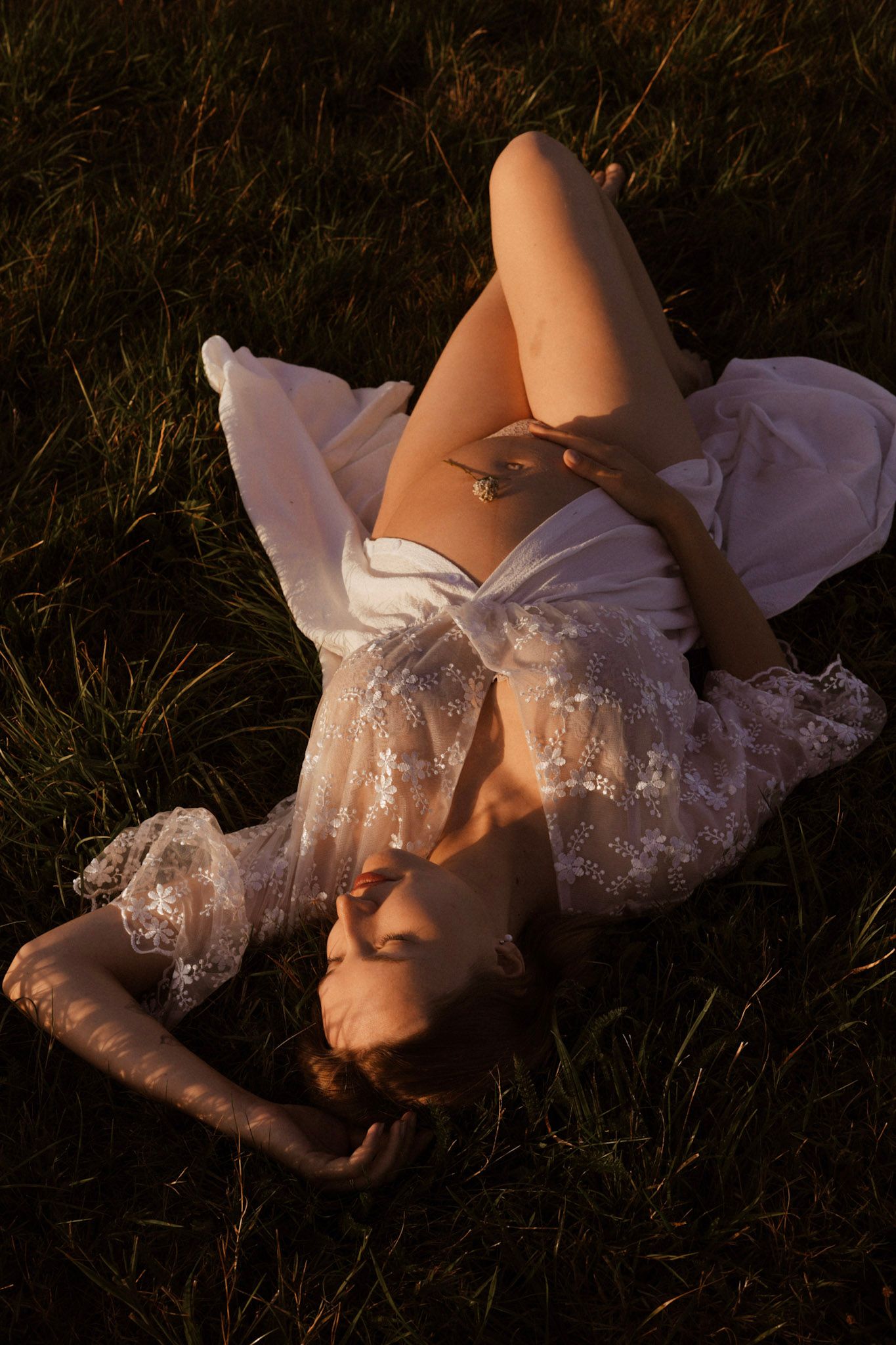 Pregnant woman in white lace robe lying on green grass in golden sunset light, gently holding her baby bump.