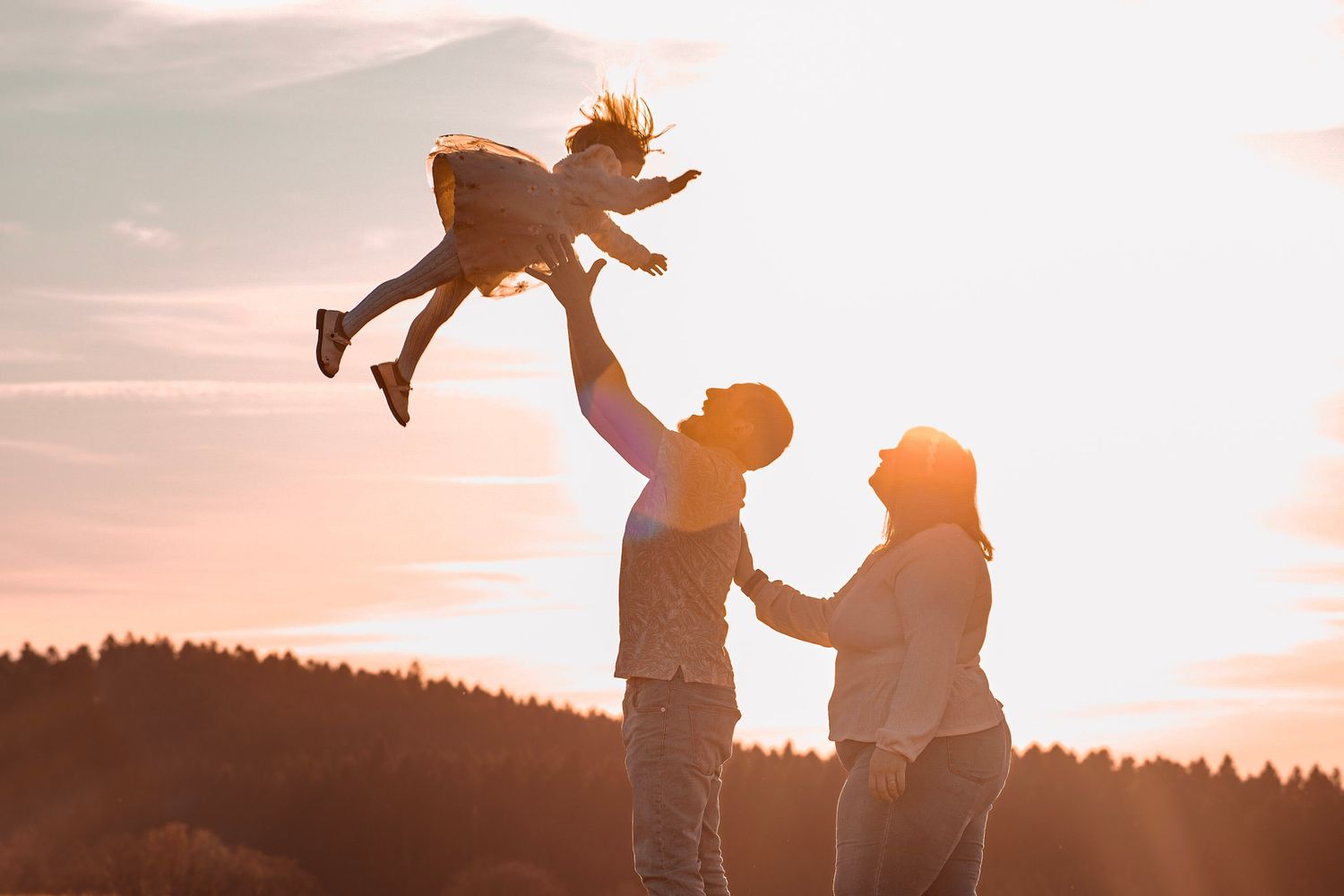 Silhouette of parents tossing child in air against golden sunset sky with rolling hills in background.