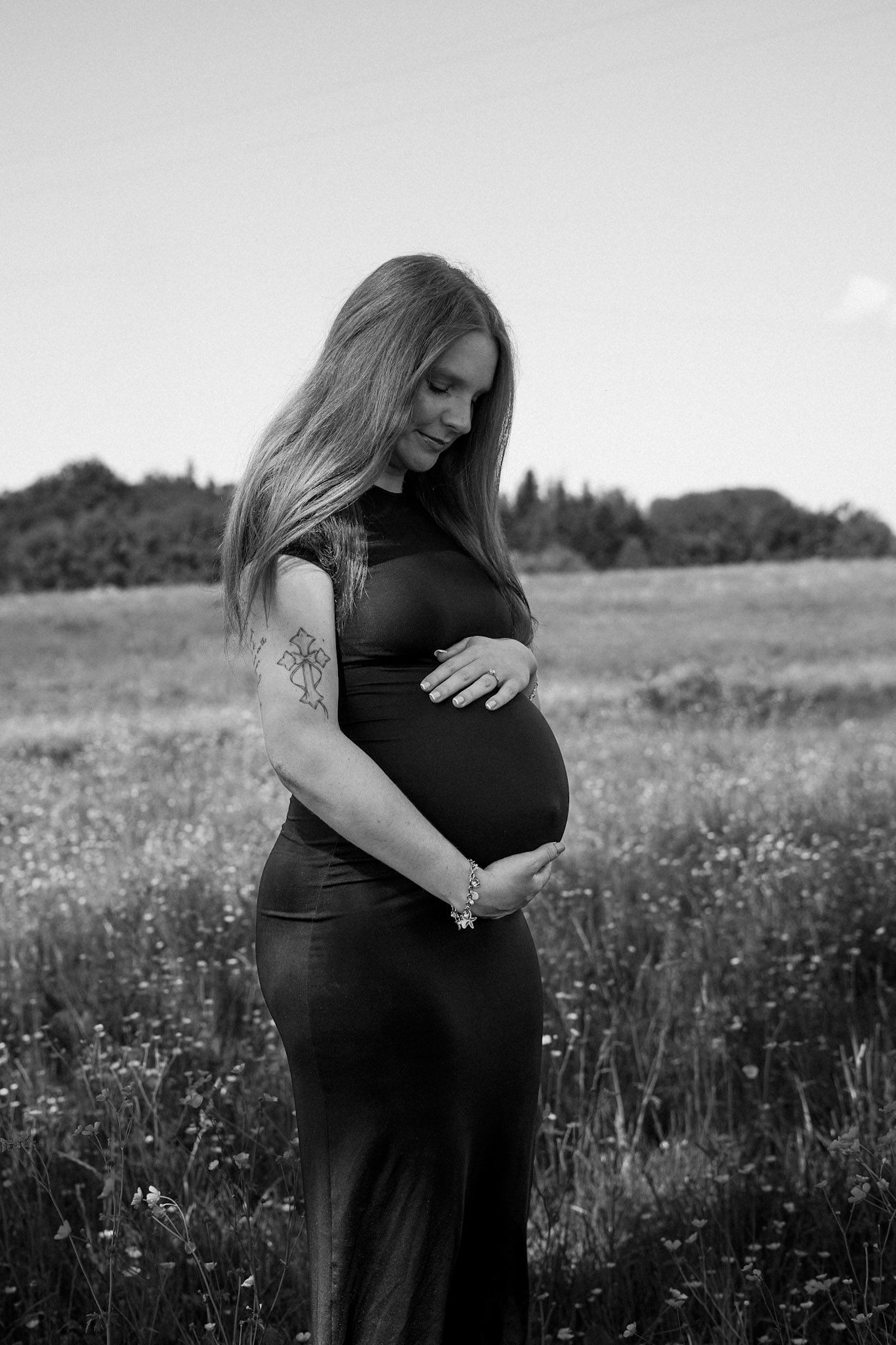 Black and white maternity photo of pregnant woman in field, cradling baby bump in elegant dress.