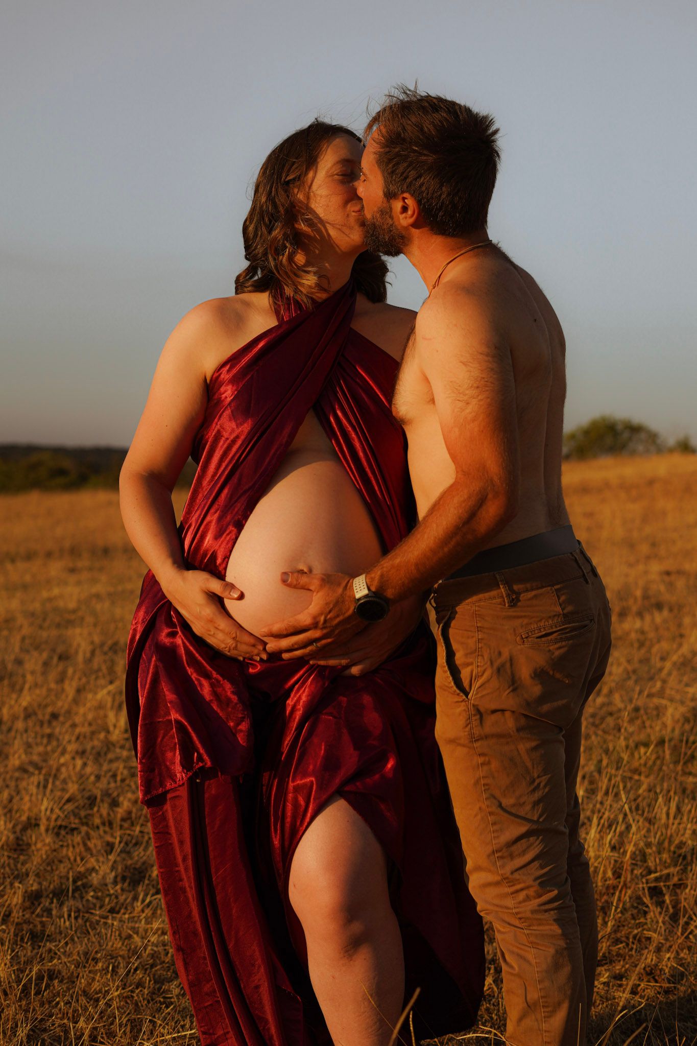 Pregnant couple kissing in golden field at sunset, man embracing woman's baby bump.