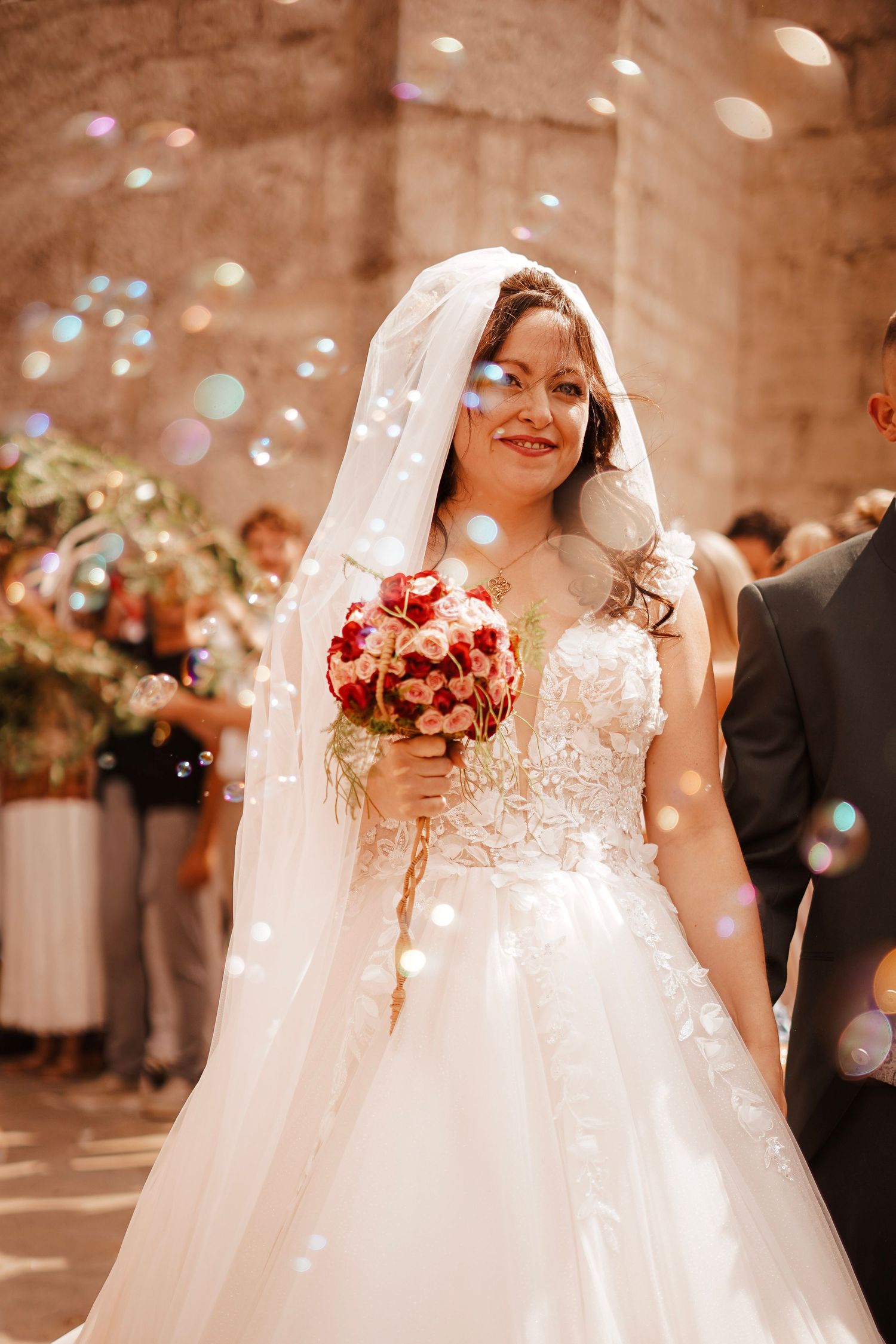 Smiling bride in white lace gown holding red bouquet surrounded by colorful soap bubbles outdoors.