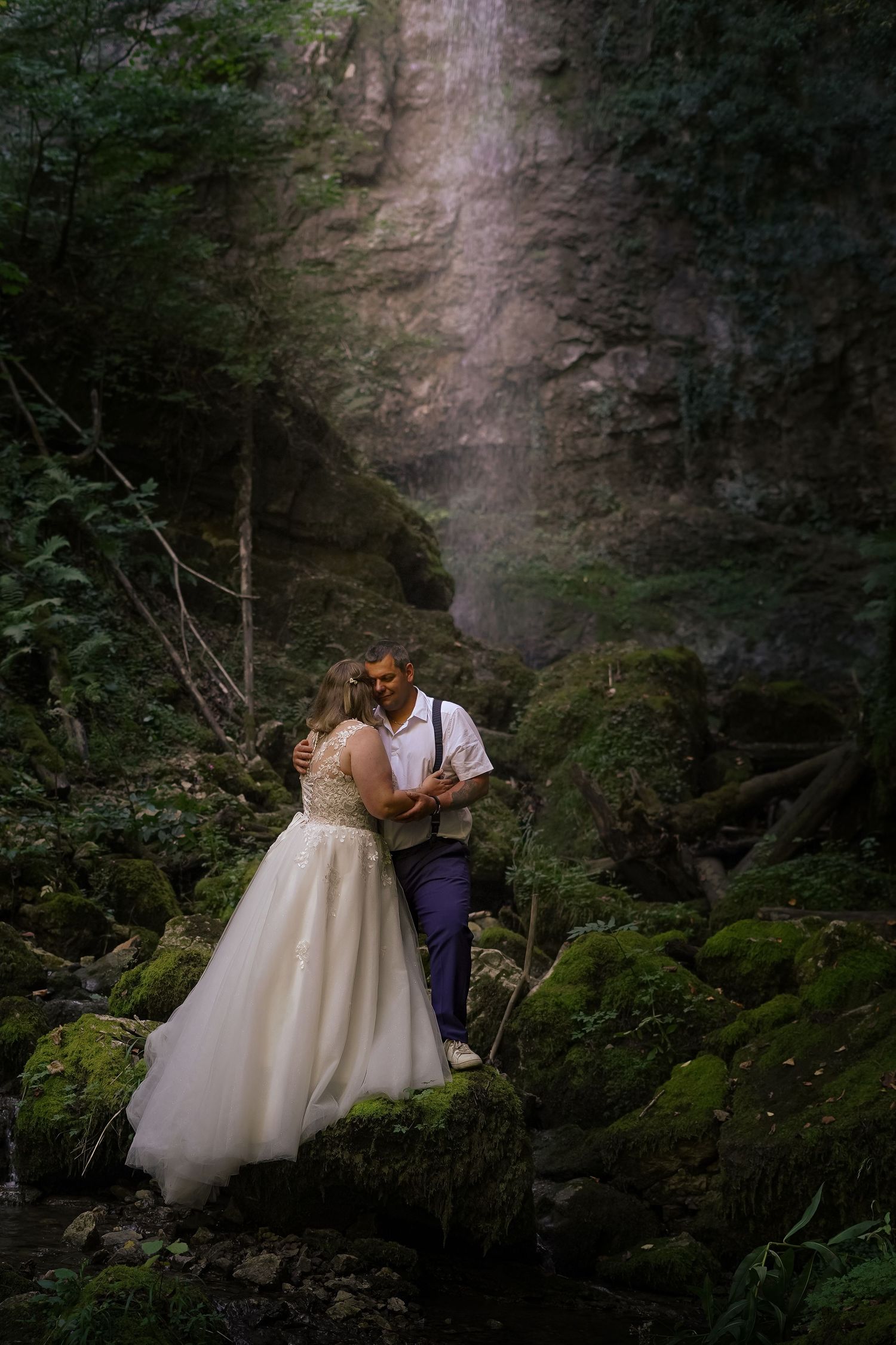 Bride and groom embracing on mossy rocks in a lush forest with a waterfall in the background.