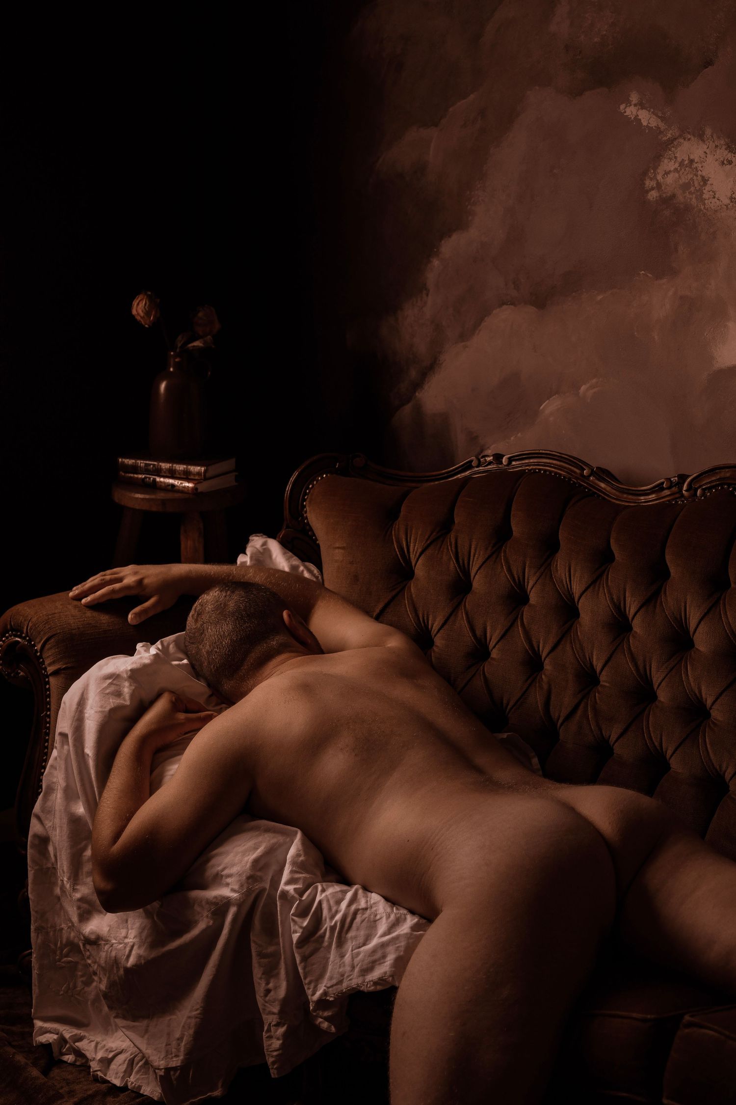 Artistic nude figure resting on a tufted velvet sofa in a dark moody room with dramatic cloudy backdrop.