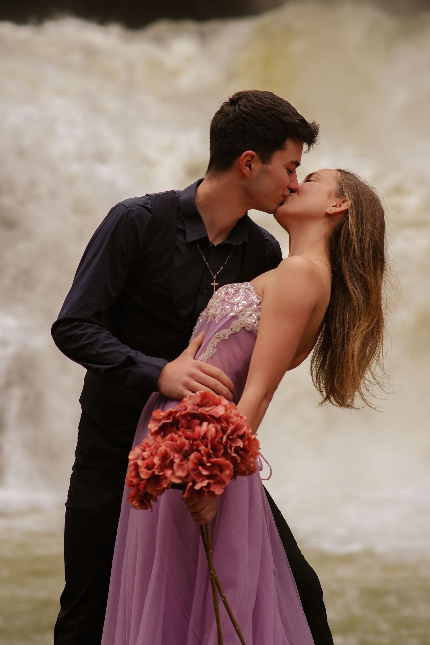 Young couple sharing a passionate kiss near a waterfall, woman holding red flowers.