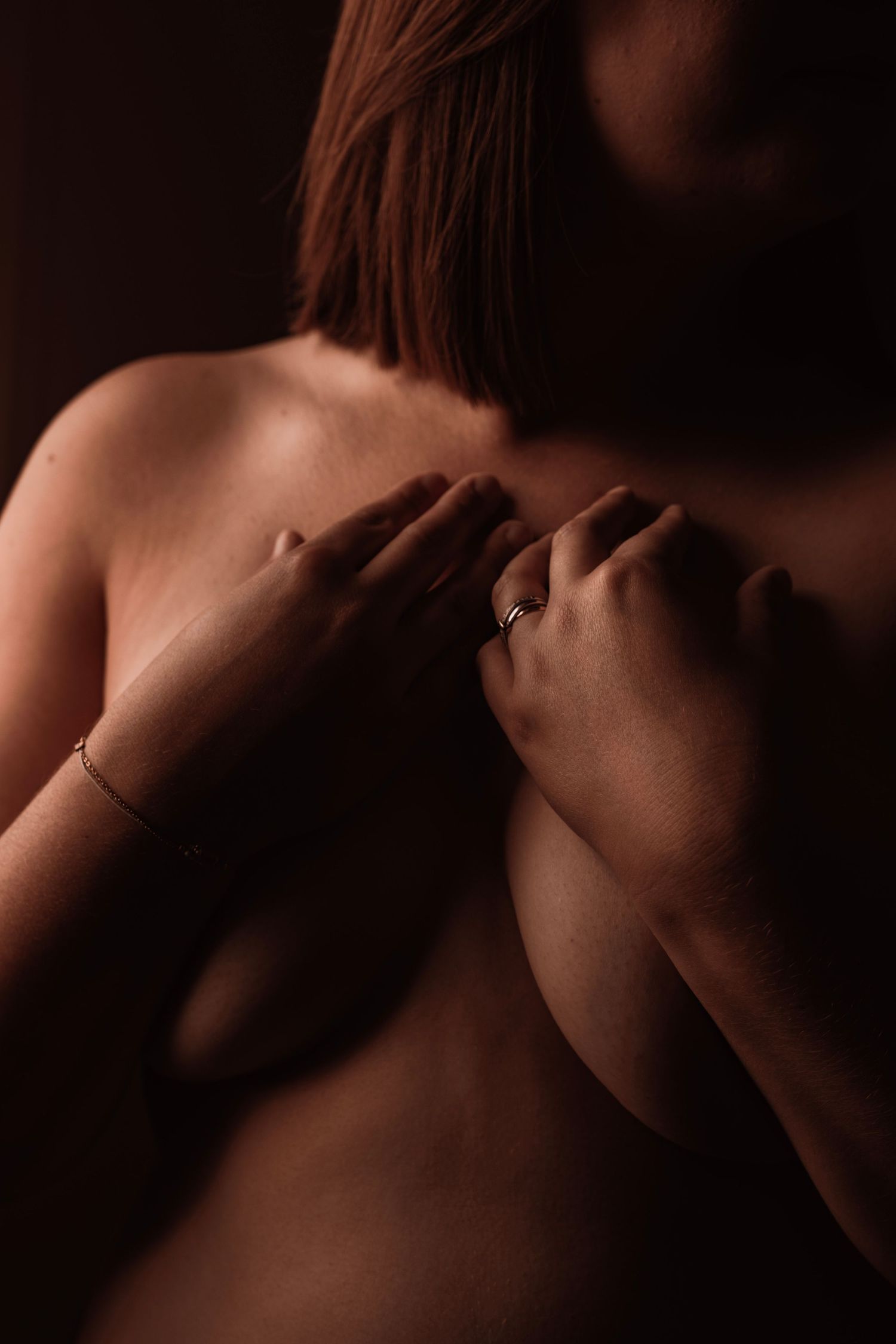 Close-up of a woman's hands with a ring and bracelet covering her chest in warm, moody low-key lighting.