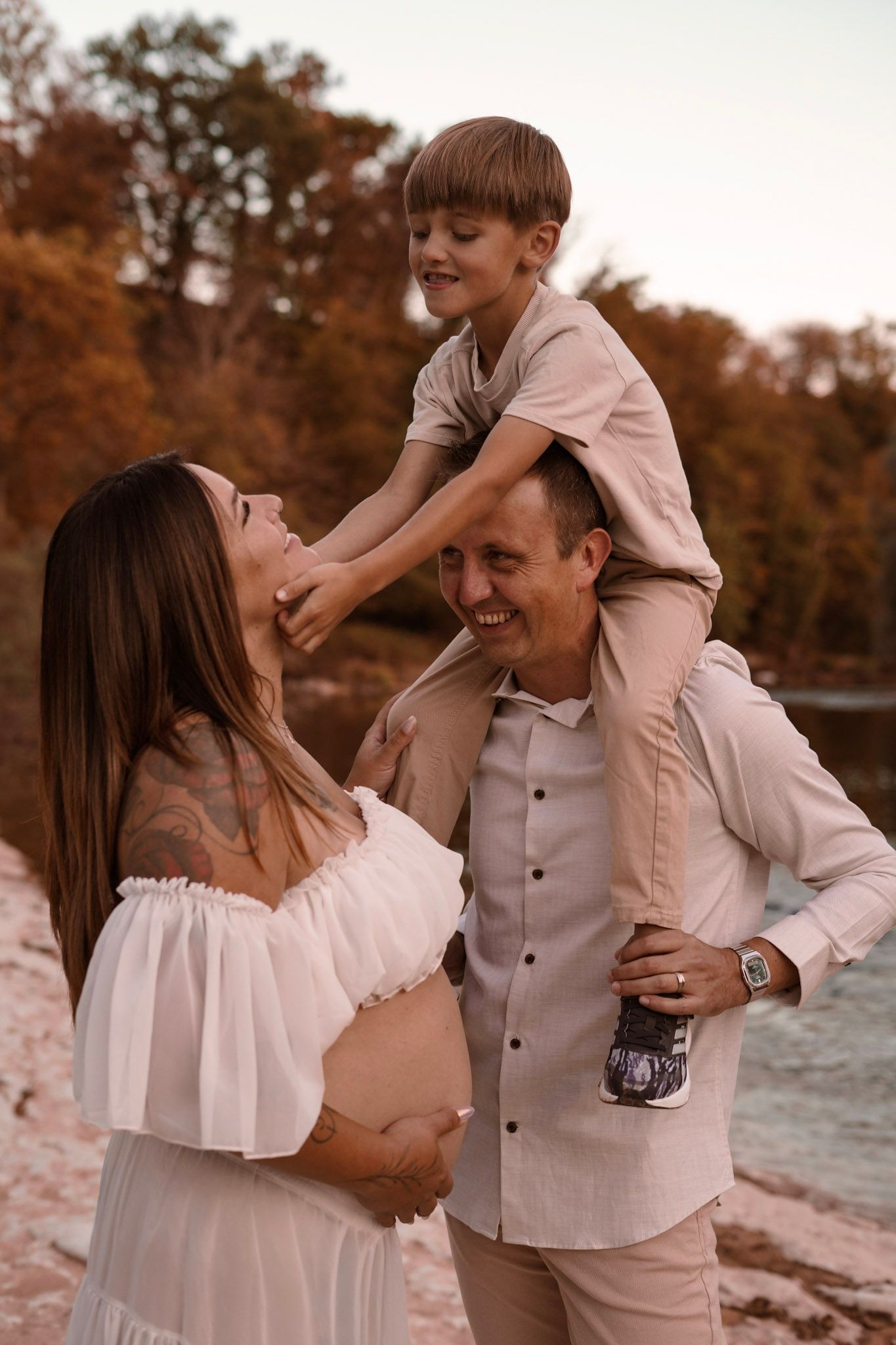 Happy family with pregnant mother, father carrying son on shoulders near autumn river.