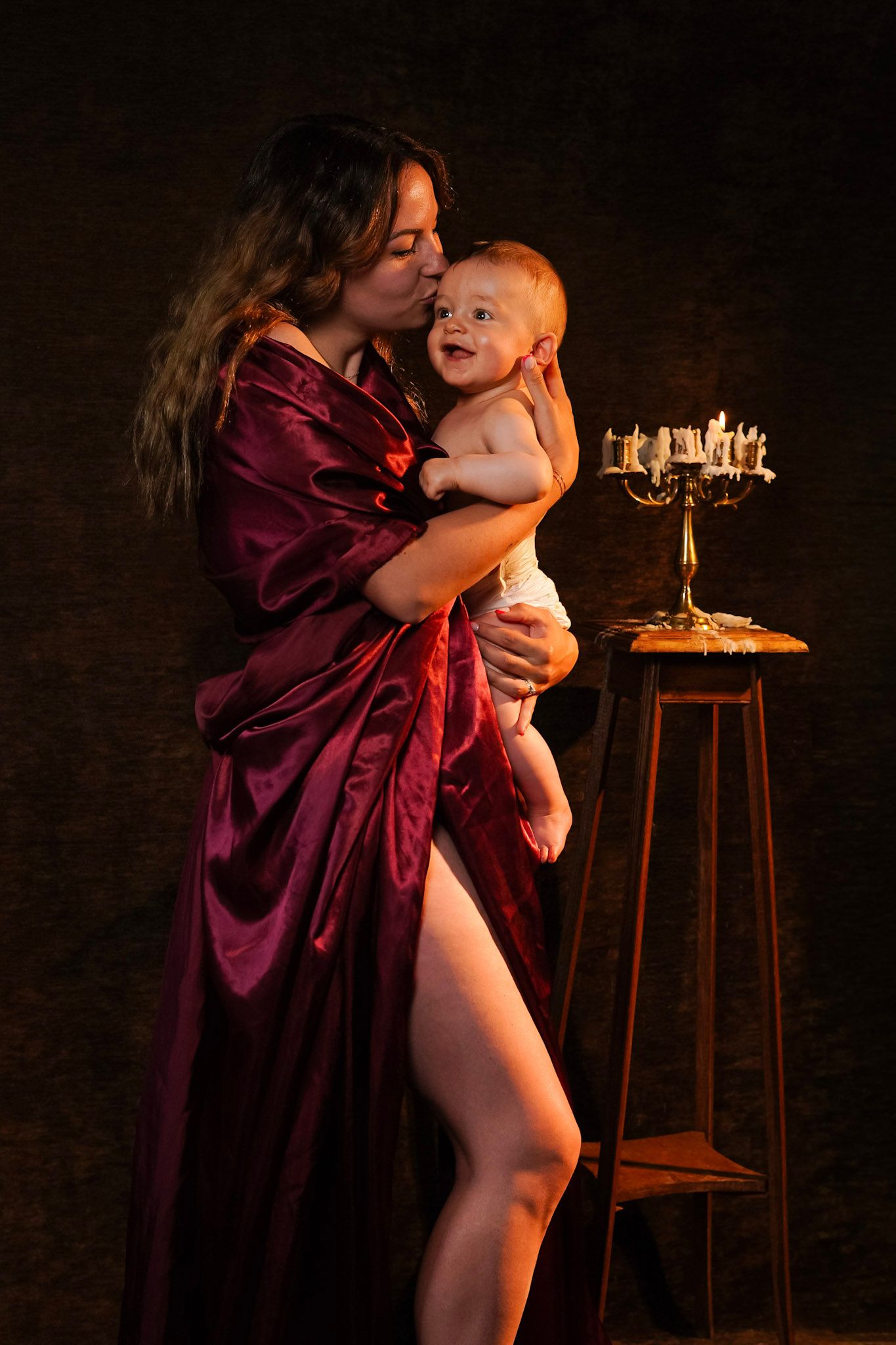 Woman in red satin robe kissing smiling baby by candlelight in dramatic dark studio portrait.