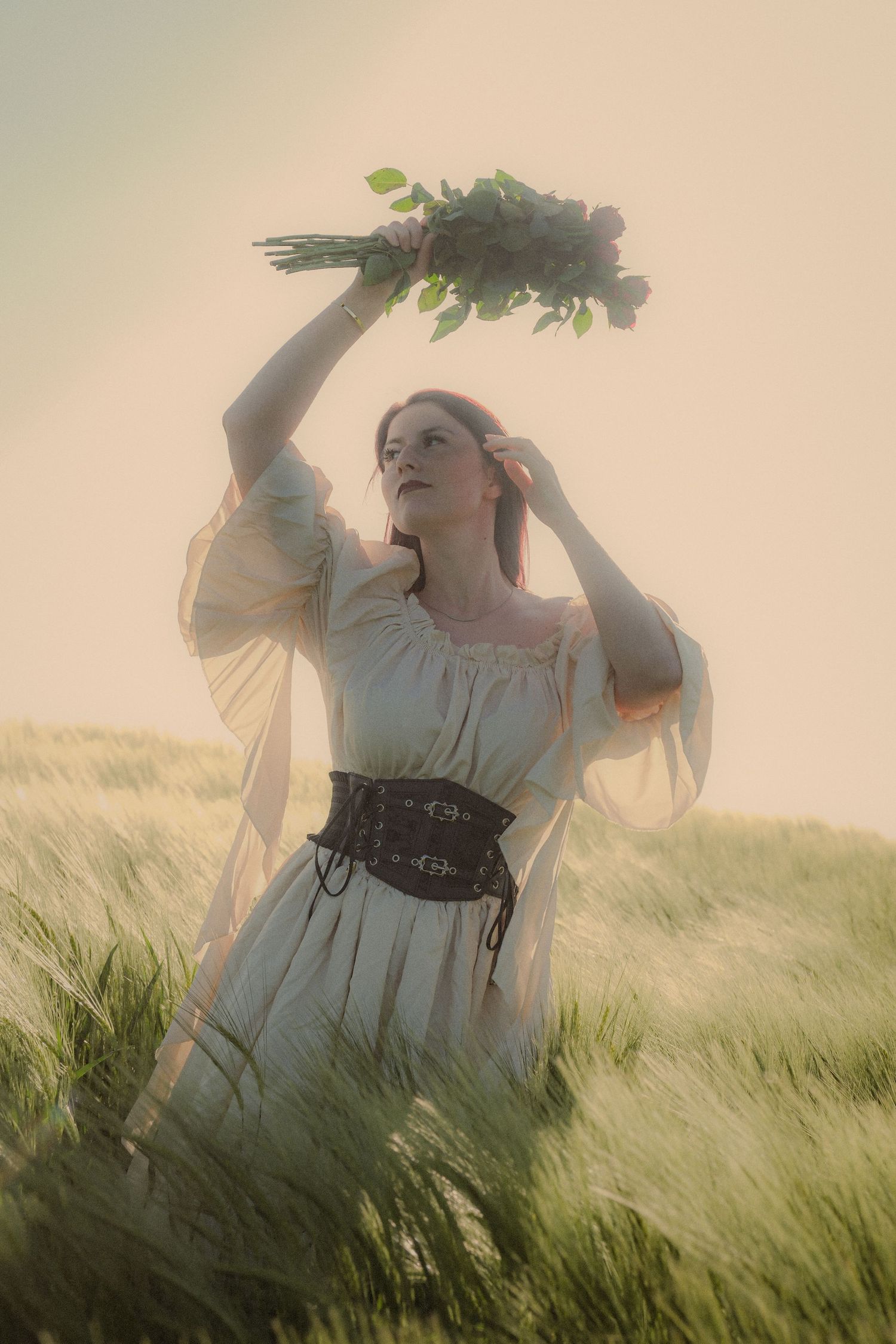 Red-haired woman in medieval dress holds wildflowers in misty wheat field.