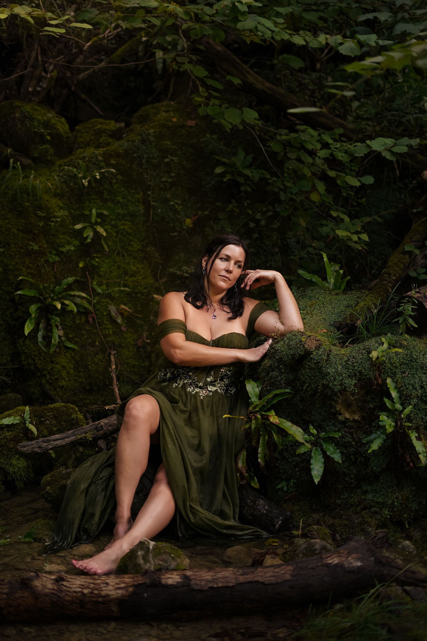 Woman in green fantasy dress sitting on mossy log in lush forest.
