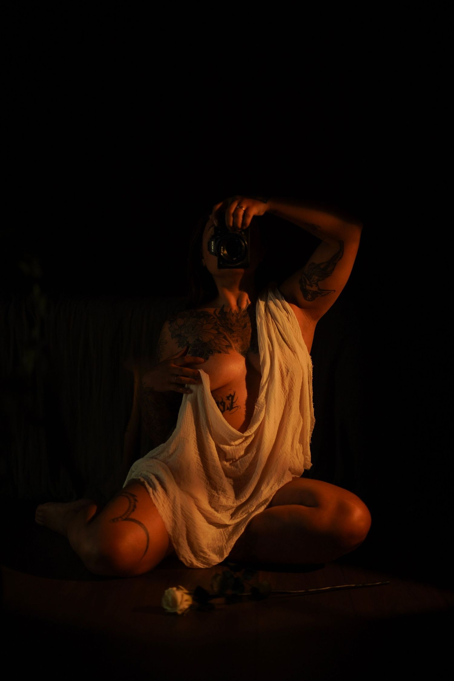 Tattooed woman in white draped fabric holding camera while sitting cross-legged in warm candlelight.