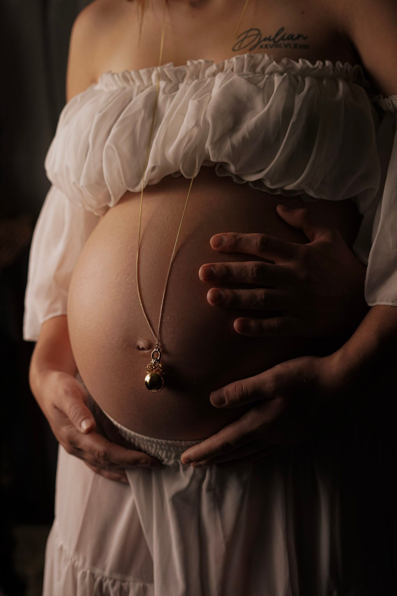 Pregnant woman in white off-shoulder dress cradling baby bump with gold necklace draped over belly.