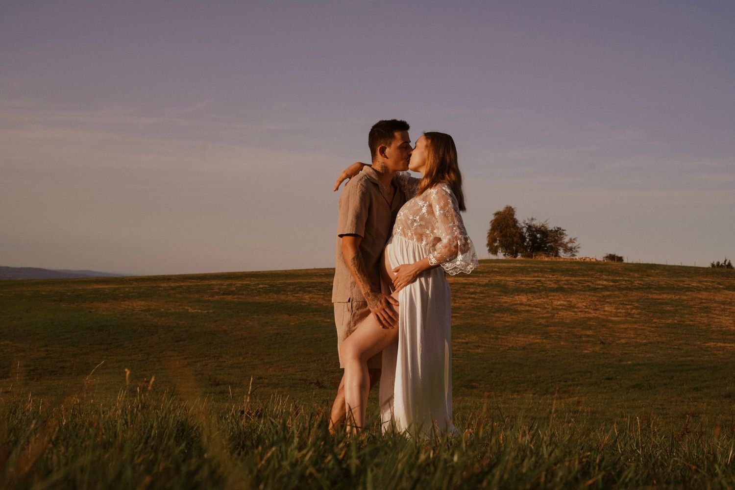 A couple shares a romantic kiss in a sunlit green field, the woman wearing a flowing white maternity dress at golden hour.