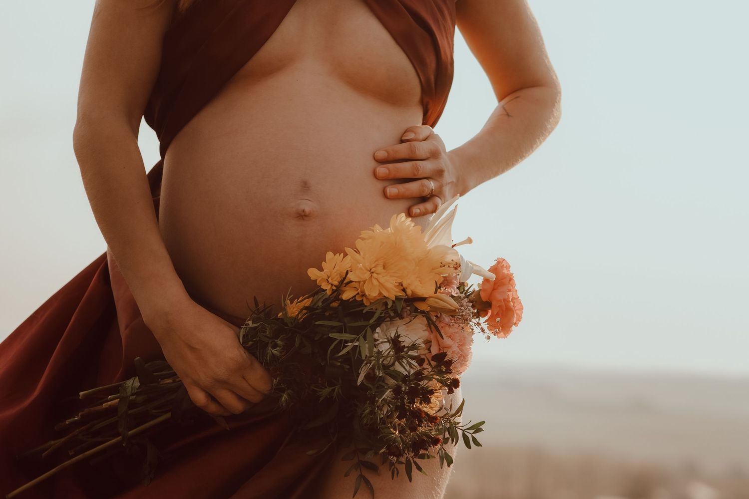 Pregnant woman in rust drape holding colorful wildflower bouquet, showcasing baby bump in soft natural light.