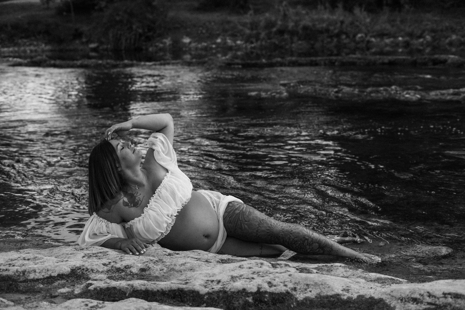 Black and white photo of a pregnant woman in white lace reclining elegantly on rocks by a river.